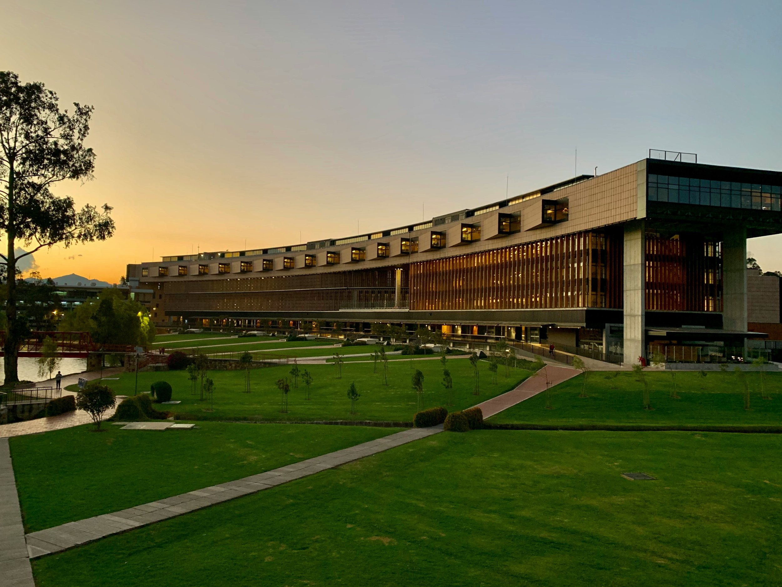 A modern building with large glass windows and a unique architectural design, surrounded by a well-maintained park with green grass, small trees, and pathways, with a sunset sky in the background.