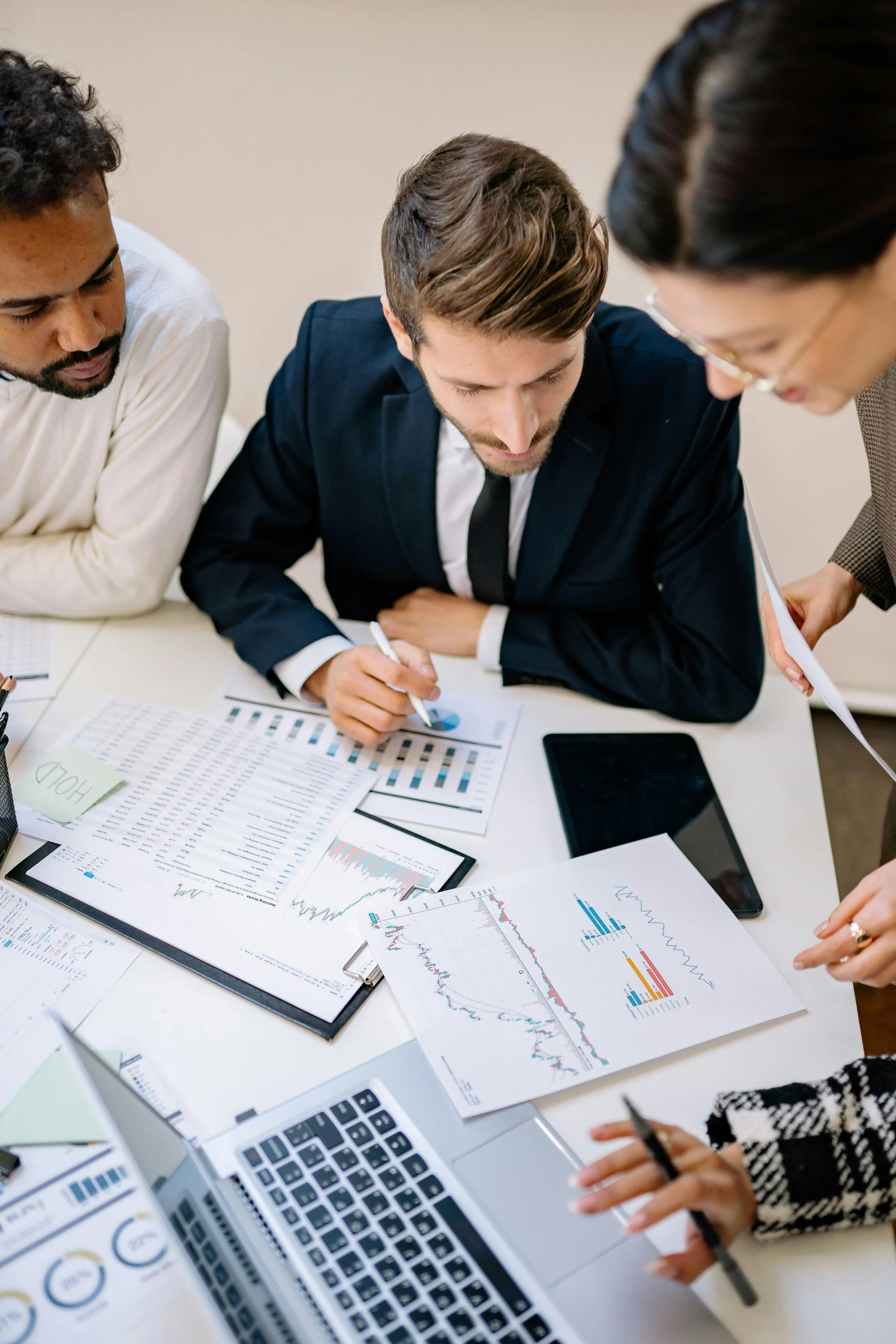 Three professionals in a business meeting reviewing financial charts and graphs spread on a table, with laptops, notebooks, and smartphones.