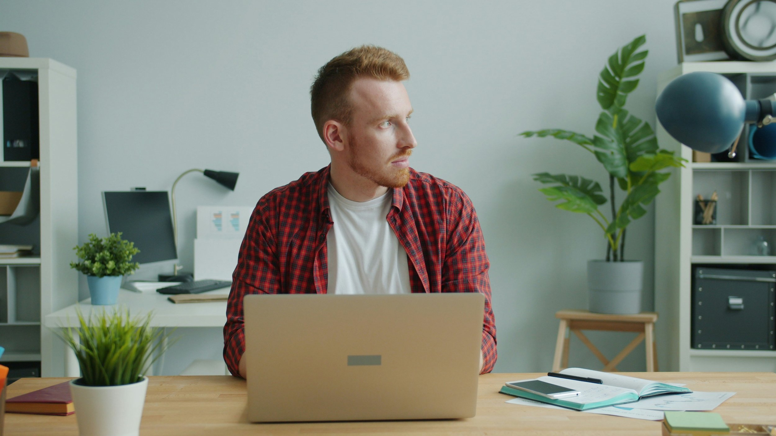 A man with red hair and a beard wearing a red plaid shirt sitting at a desk in a modern office space, looking to his left. The desk has a laptop, notebooks, a smartphone, and a potted plant. The background features shelving with books, plants, and office equipment.