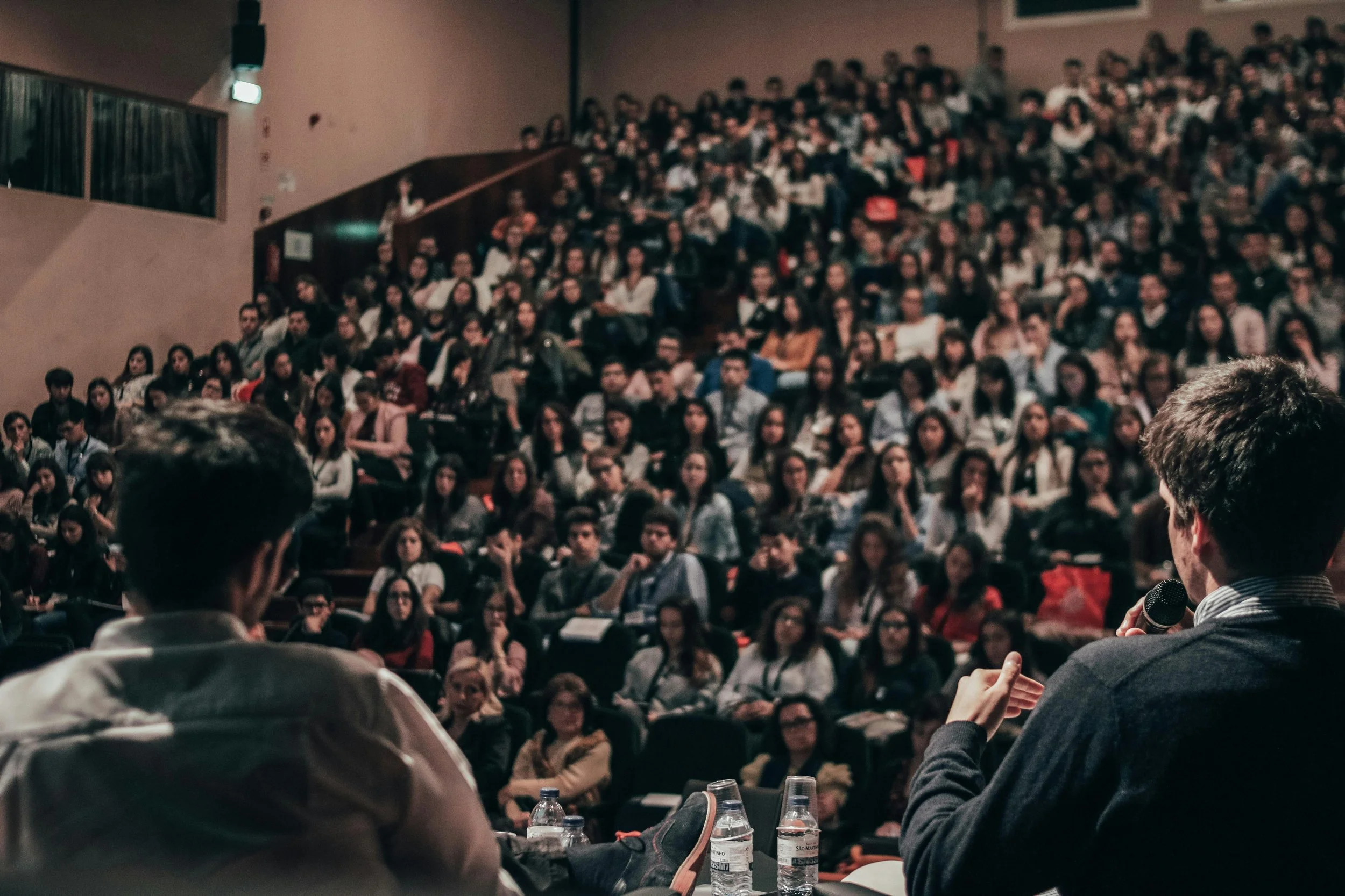 A large audience seated in theater seats during a presentation or lecture, with a person speaking into a microphone in the foreground.