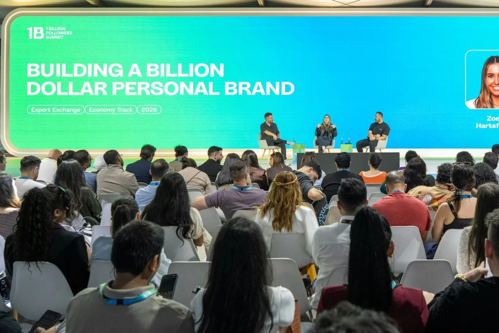 A large conference room filled with a diverse audience attending a presentation on stage, where three speakers are seated and a large screen displays a green and blue background with the words 'Building a Billion Dollar Personal Brand'.
