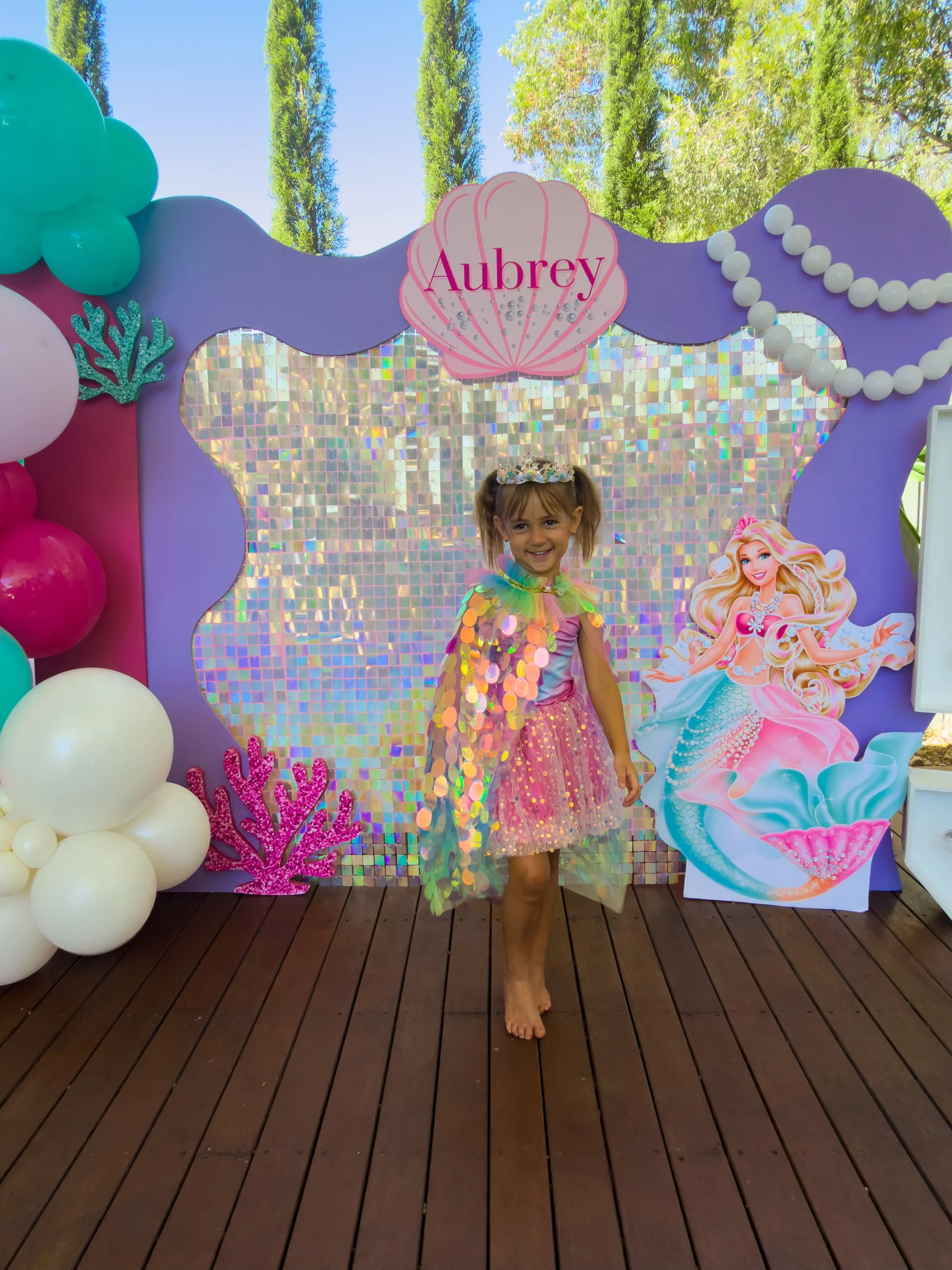 A young girl dressed as a mermaid standing on a wooden deck at a birthday party with an under-the-sea themed backdrop, balloons, and a large cutout of a mermaid.