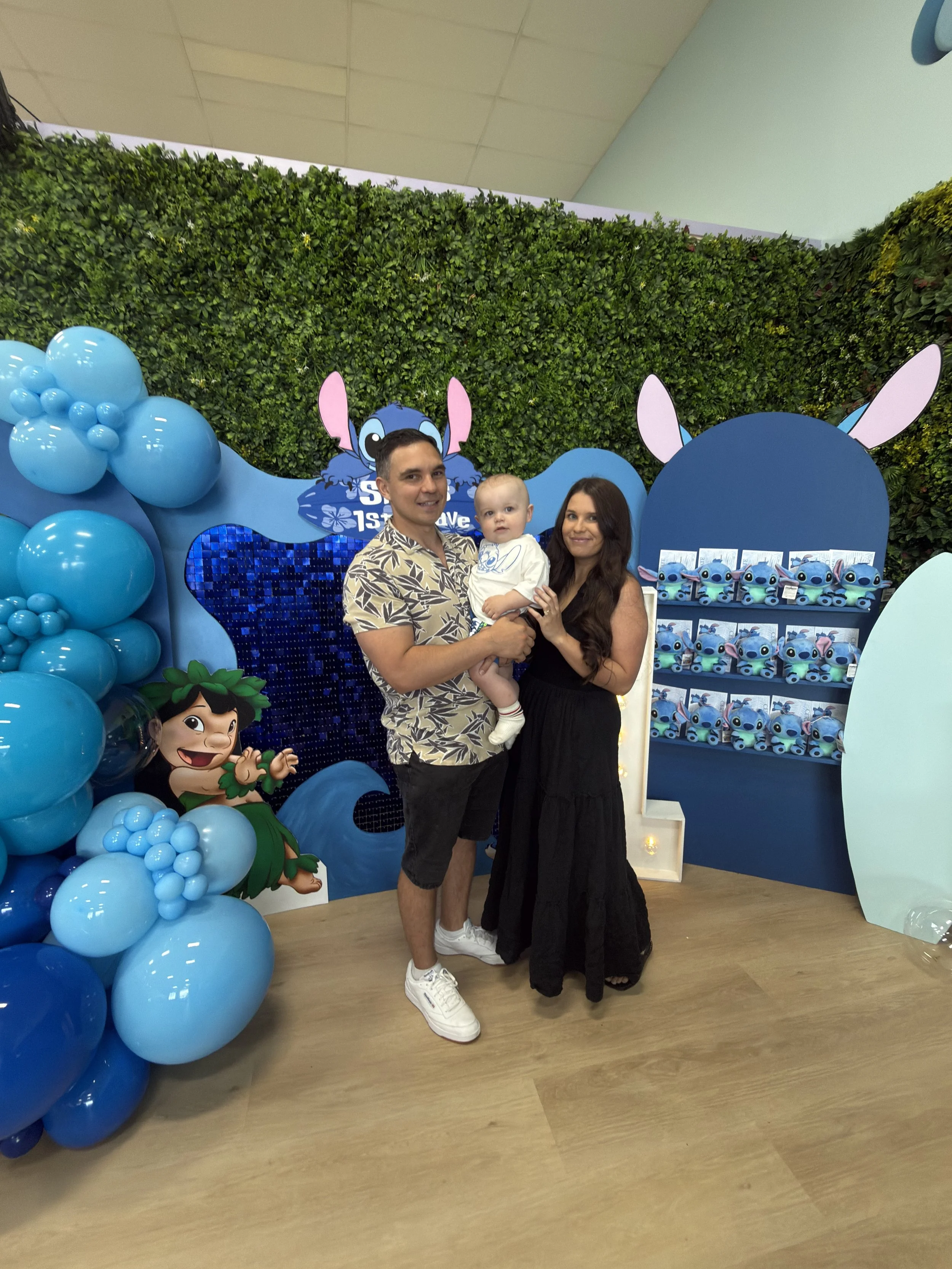 A family of three standing in front of a Disney's Lilo & Stitch themed backdrop with blue balloons and character cutouts, at a birthday celebration.