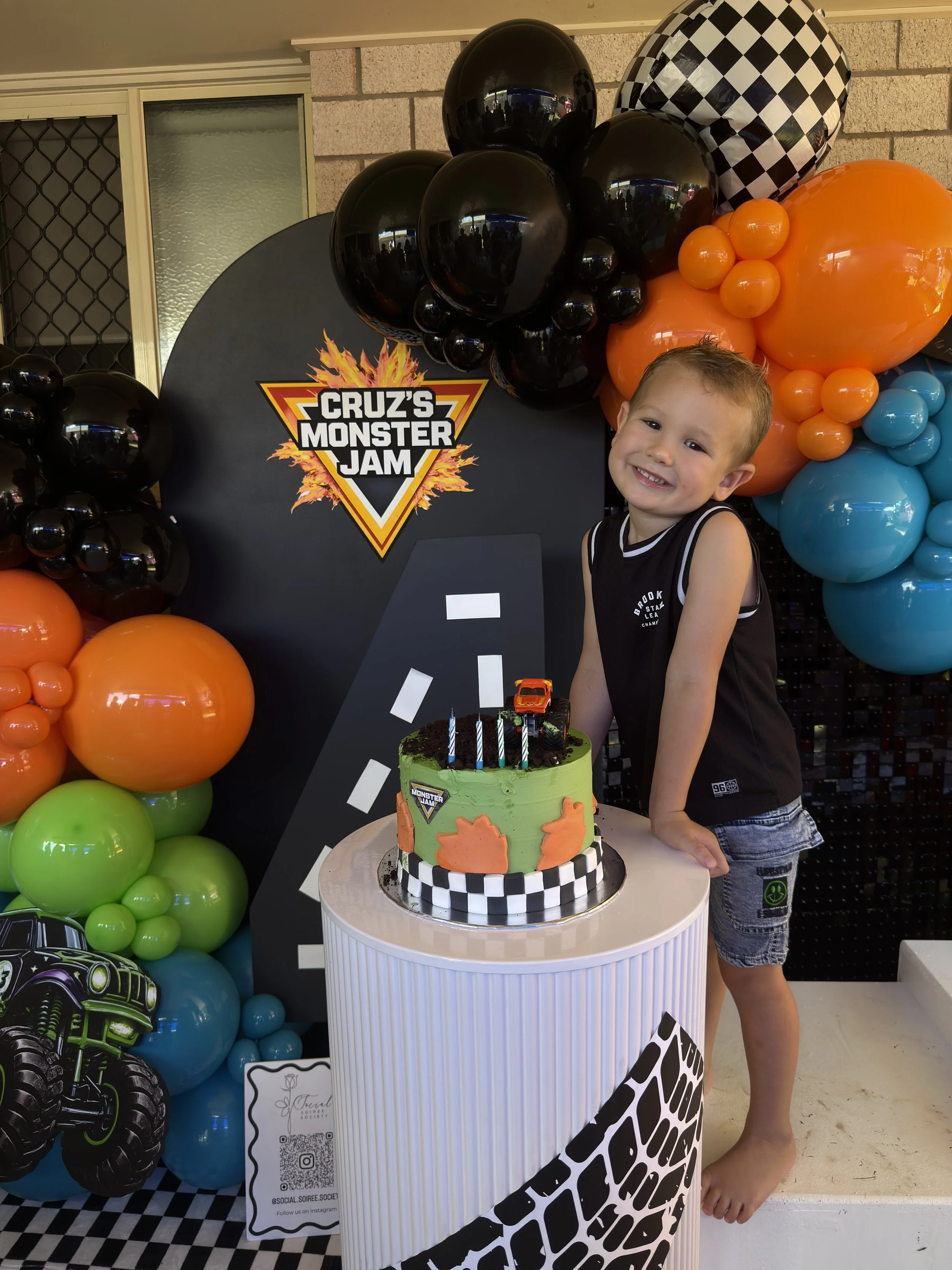 A boy standing next to a birthday cake decorated with a monster jam theme, surrounded by colorful balloons, with a backdrop featuring a Monster Jam logo and checkered and flame patterns.