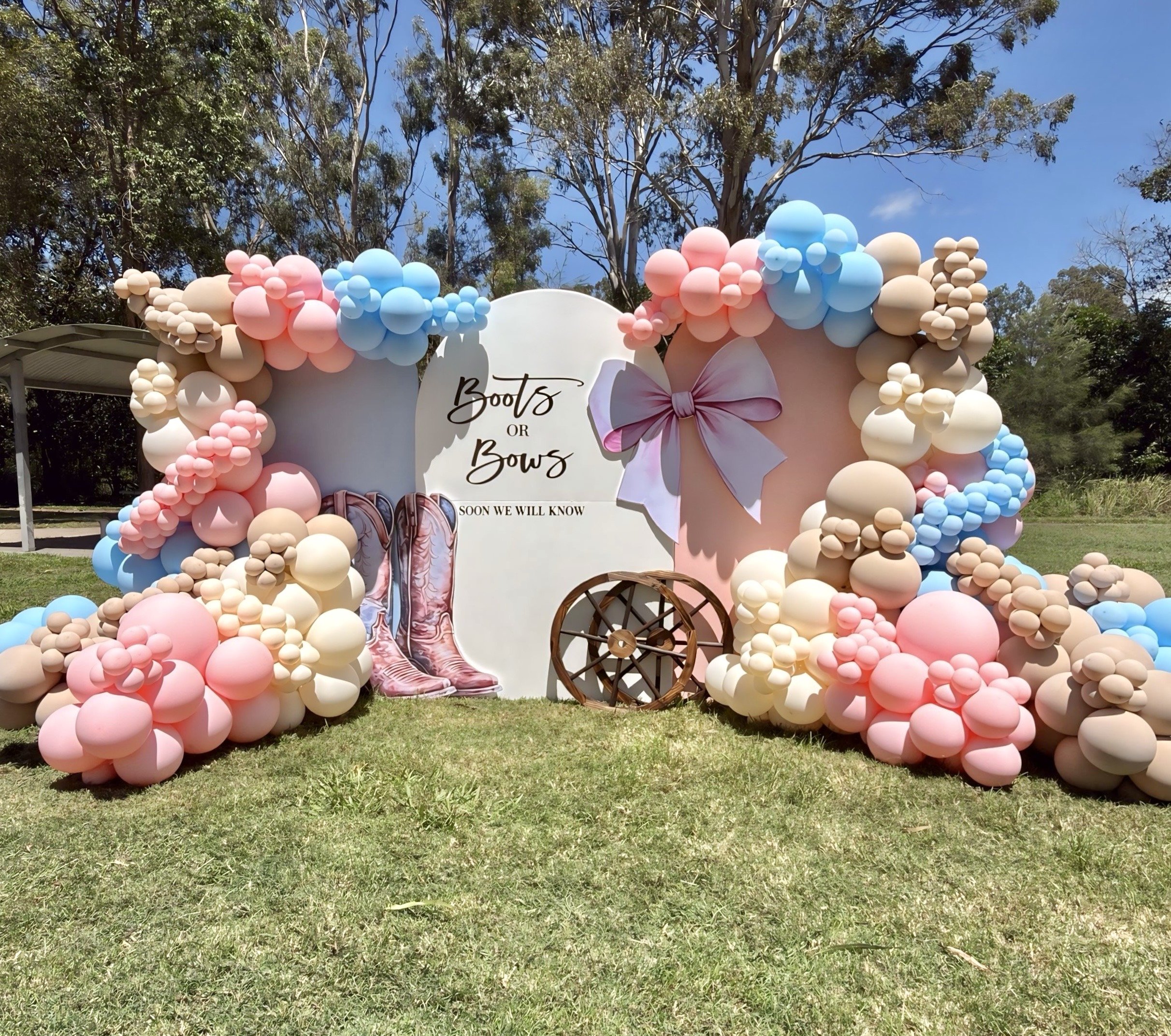 Decorative balloon arch in pink, blue, and beige tones at an outdoor event, with a sign reading "Boots or Bows" and illustrations of childrens shoes and a bow, set against a backdrop of trees and a blue sky.