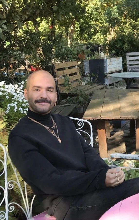 A man with a mustache and goatee, wearing a black turtleneck and gold jewelry, sitting on a white wrought iron chair outdoors in a garden with trees, plants, and wooden furniture.