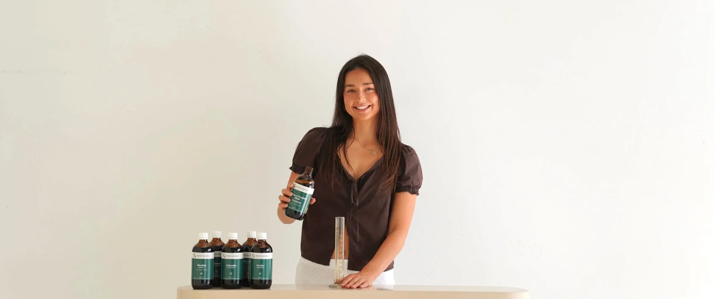 A young woman with long dark hair smiling while holding a bottle of medicine or supplement, standing behind a table with five similar bottles of herbal medicine and a measuring cylinder in a plain setting.