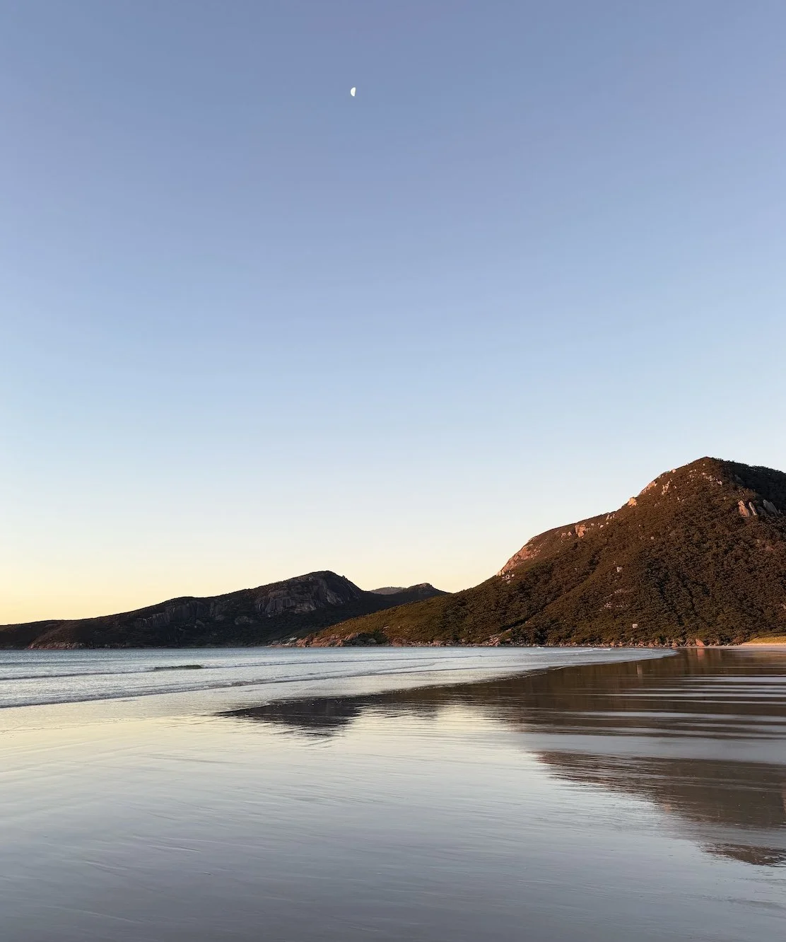 Sunset over calm beach with mountains in the background and the moon visible in the sky