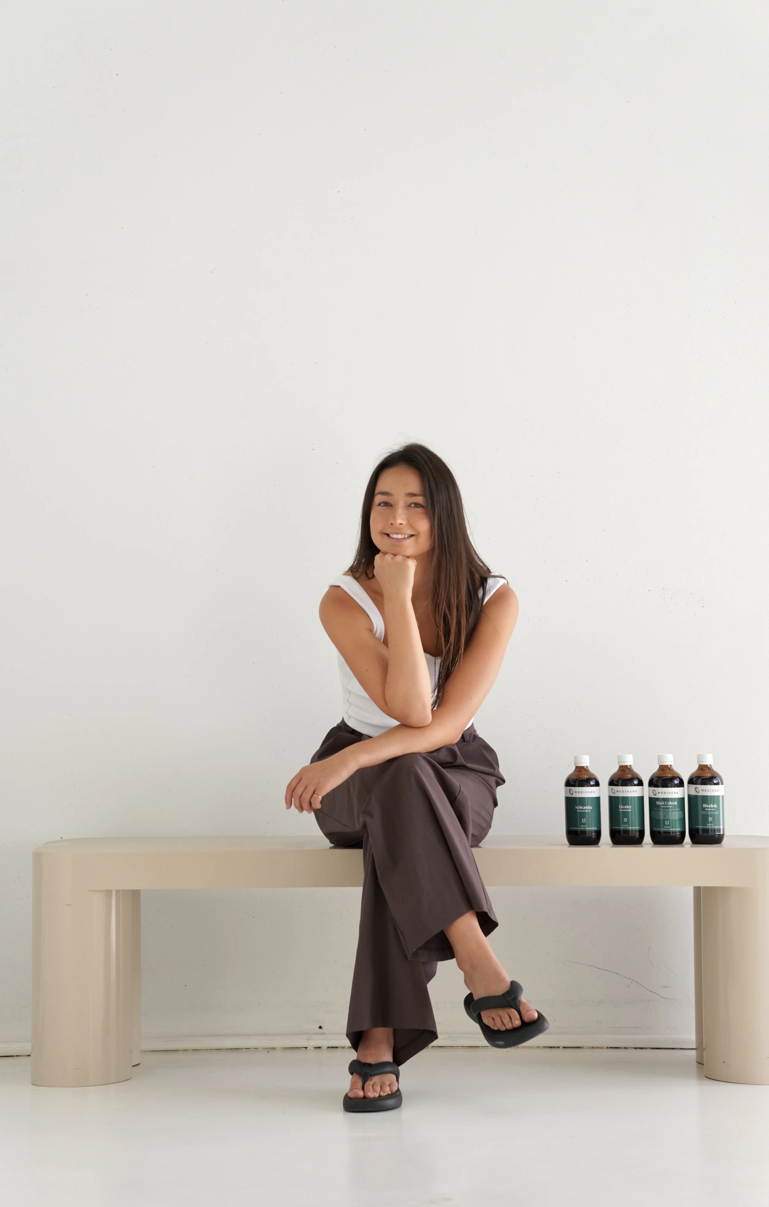 A woman sitting on a beige bench with her chin resting on her hand, smiling at the camera. She is wearing a white sleeveless top, brown pants, and black sandals. Four bottles with black labels are placed on the bench beside her, against a plain white wall.