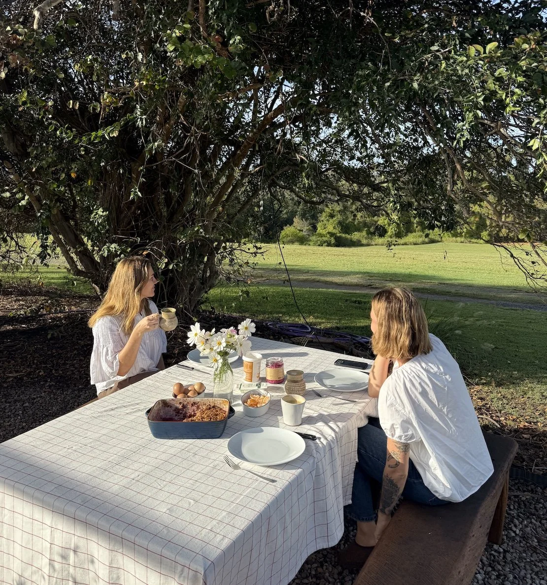 Two women having breakfast outdoors at a table under a large tree, with a grassy field in the background, on a sunny day.