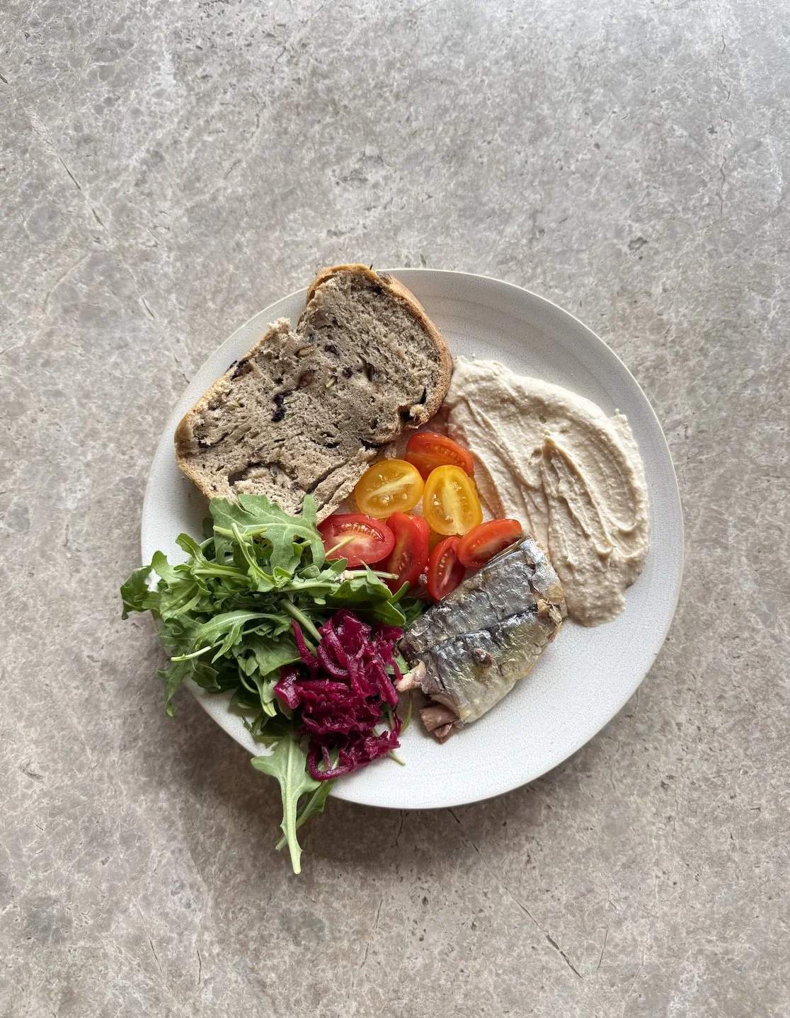 Plate of food with sourdough bread, spread of creamy hummus, cherry tomatoes, sardines, rocket, and shredded purple beetroot, on a light gray surface.