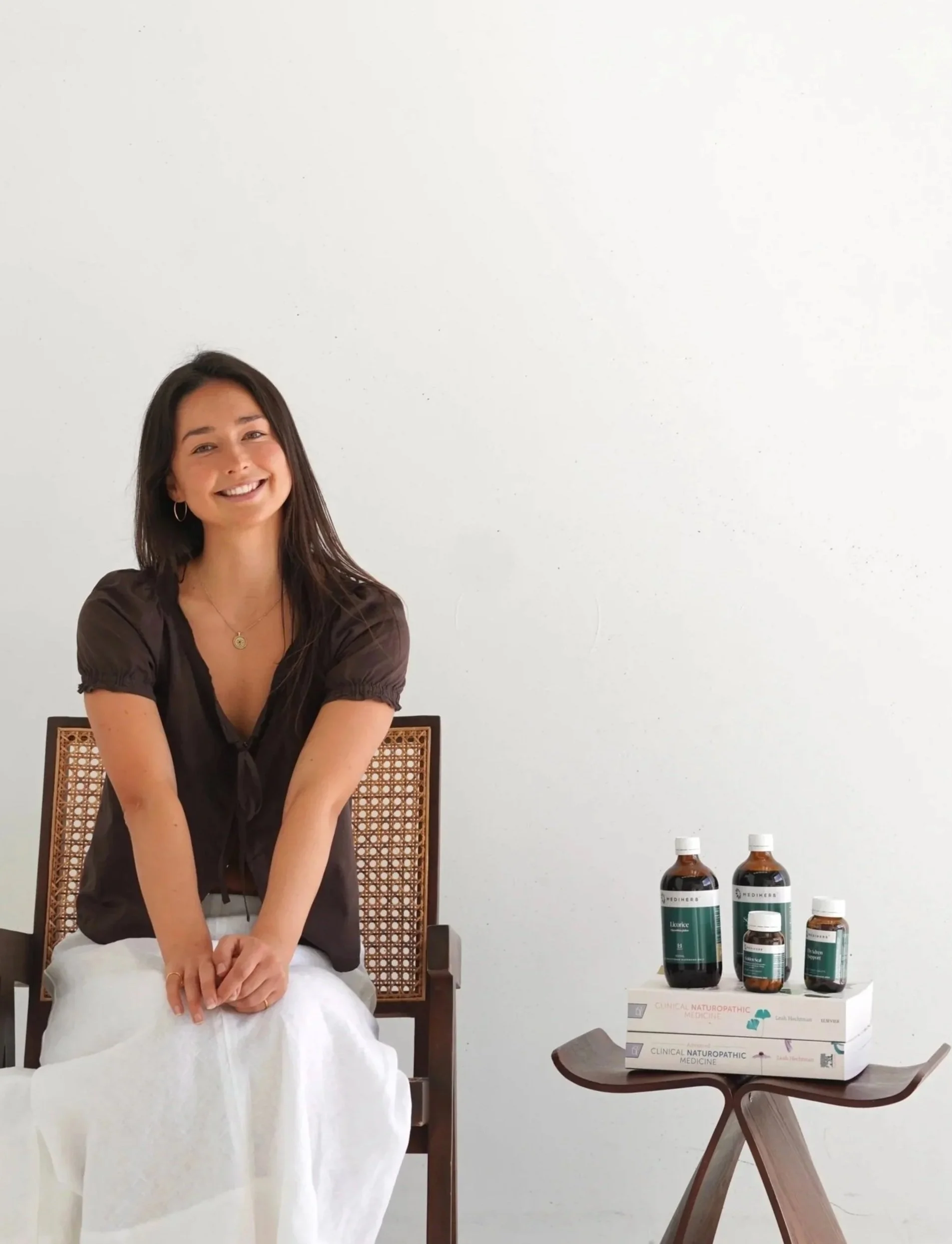 A woman with dark hair and a smile, sitting on a wooden chair, wearing a dark blouse and a white cloth on her lap, with bottles of health products on a small table beside her, in front of a plain white wall.