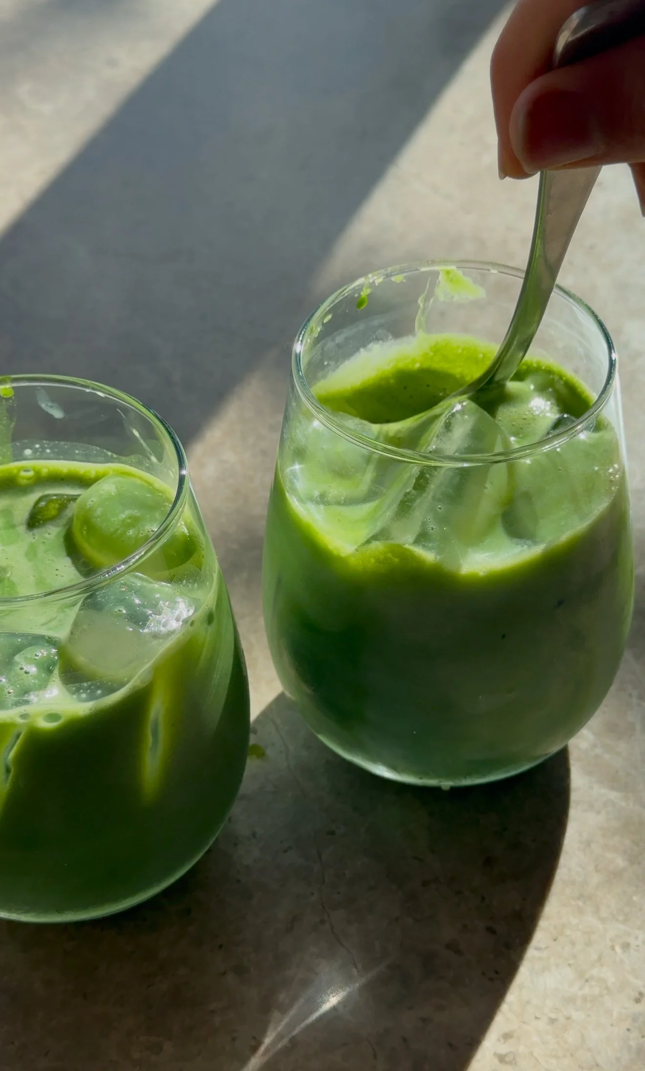 Two glasses of iced matcha, one being stirred with a spoon, on a gray surface with sunlight.