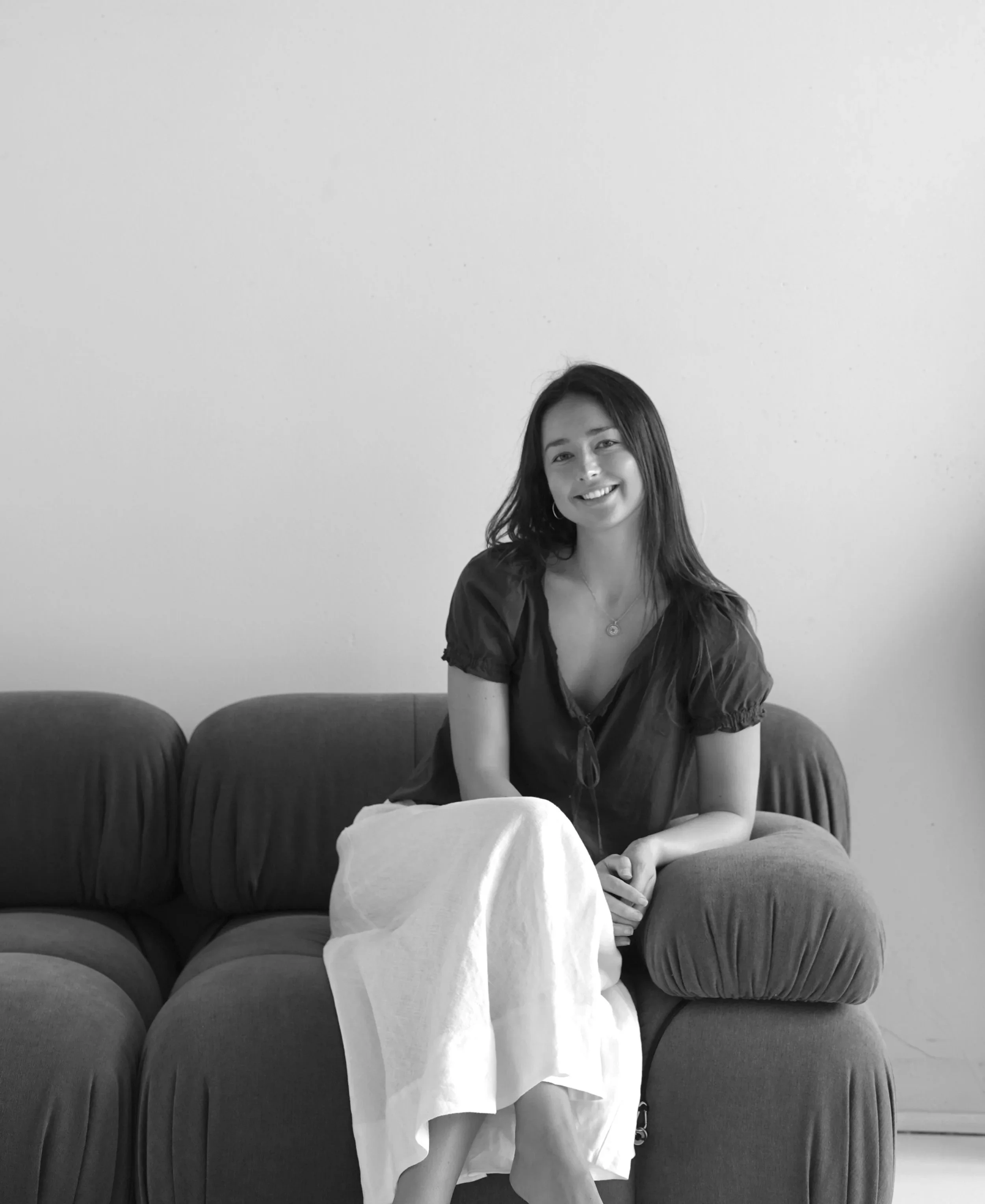 A young woman with long dark hair, wearing a V-neck top, sitting on a sofa with ottoman and smiling at the camera, in front of a blank wall.