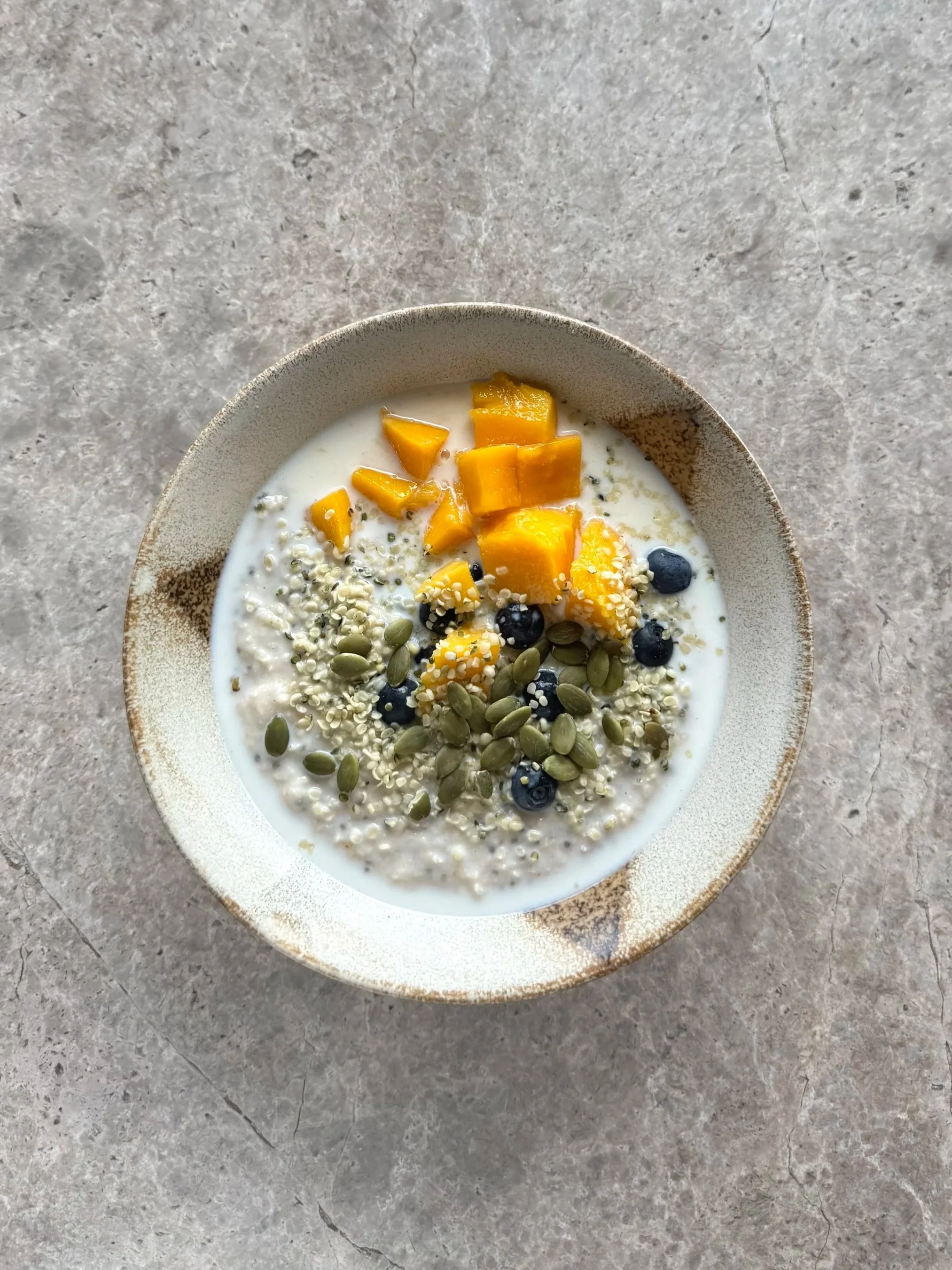 A ceramic bowl filled with a oatmeal, yogurt, topped with chopped mango, blueberries, pumpkin seeds, hemp seeds, and sesame seeds, on a gray textured surface.