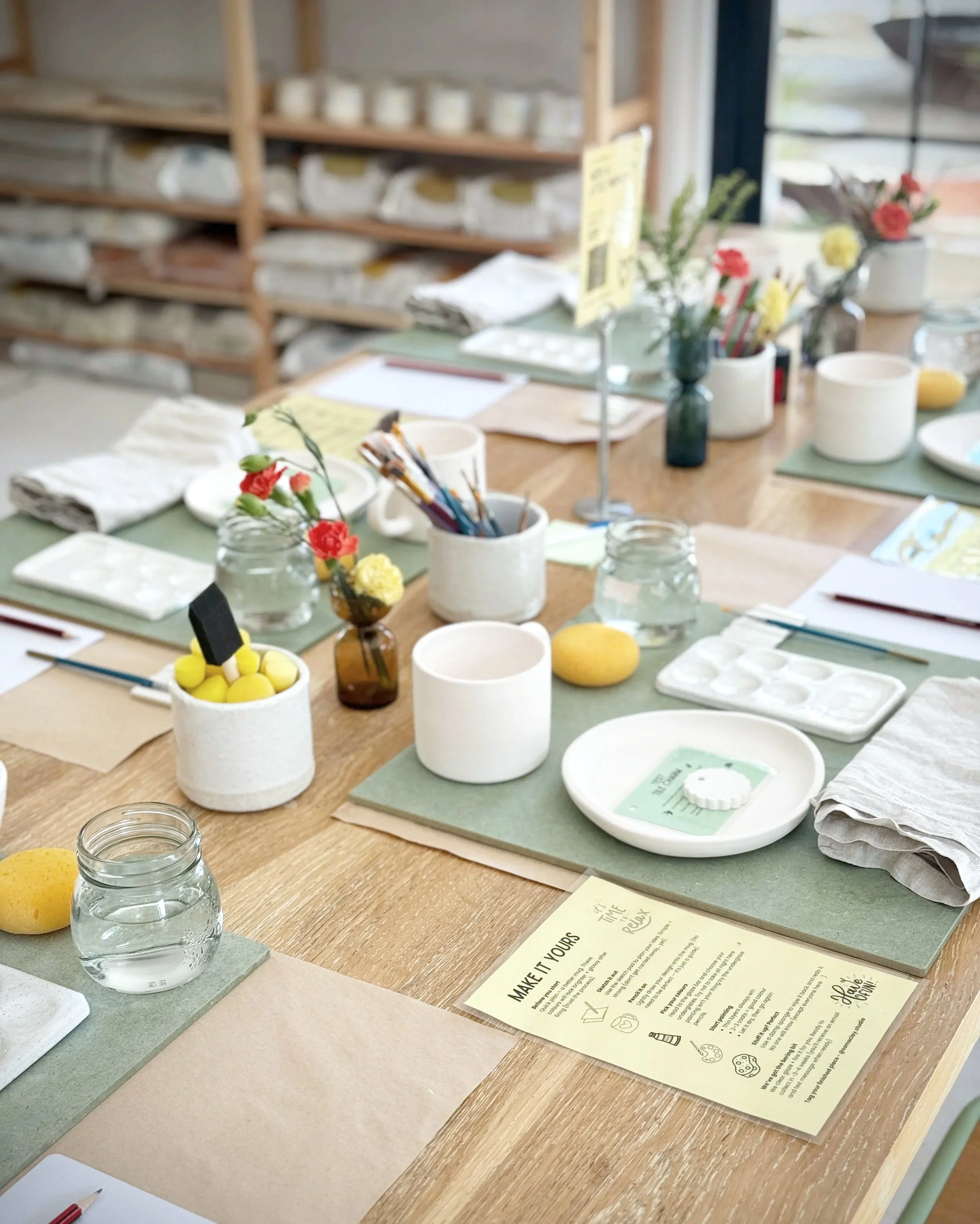 A crafting or DIY workspace with various supplies on a wooden table, including jars of water, flowers, brushes, a palette, paper, and informational sheets, with shelves filled with containers in the background.