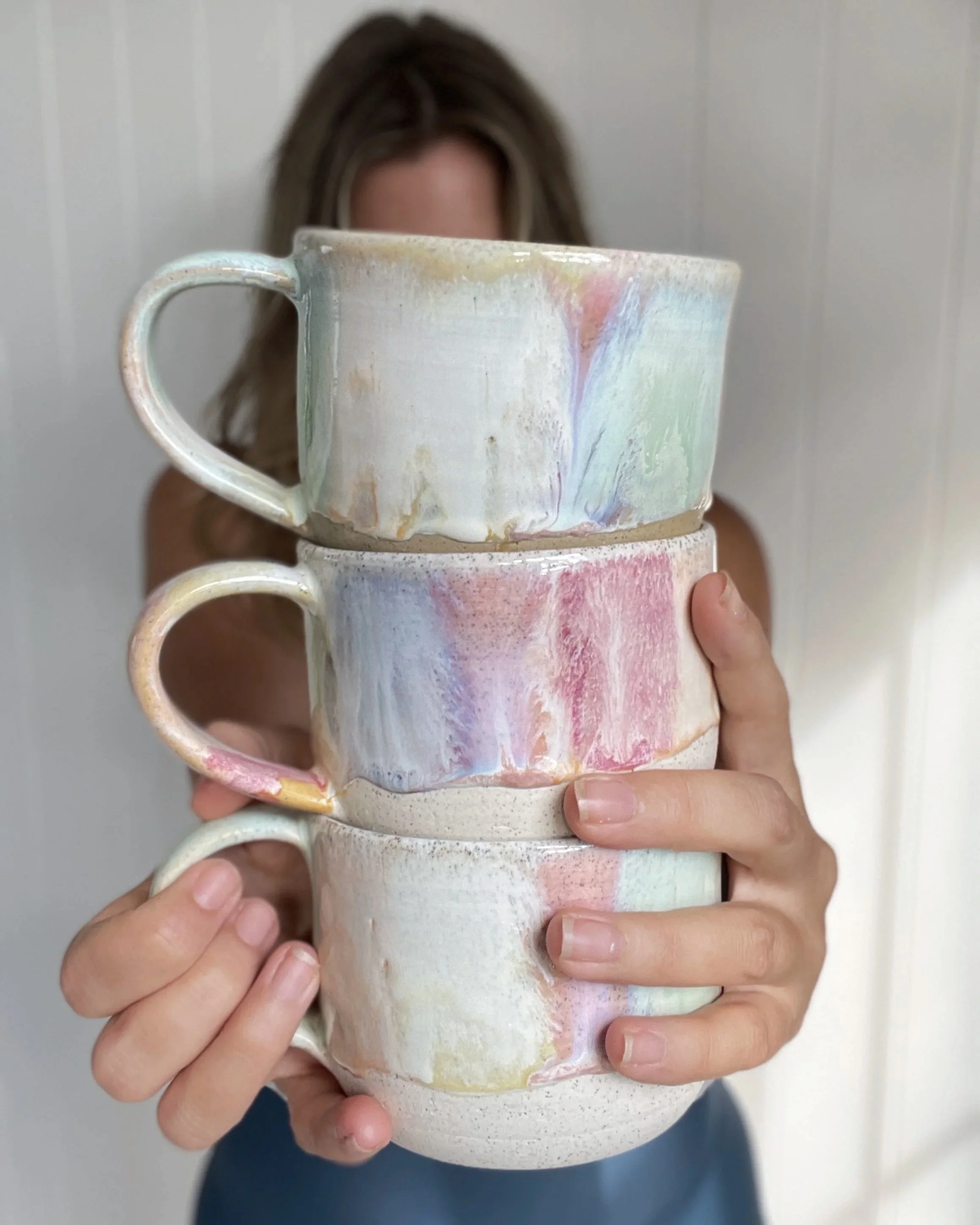 Person holding a stack of colorful, glazed ceramic mugs against a plain background.