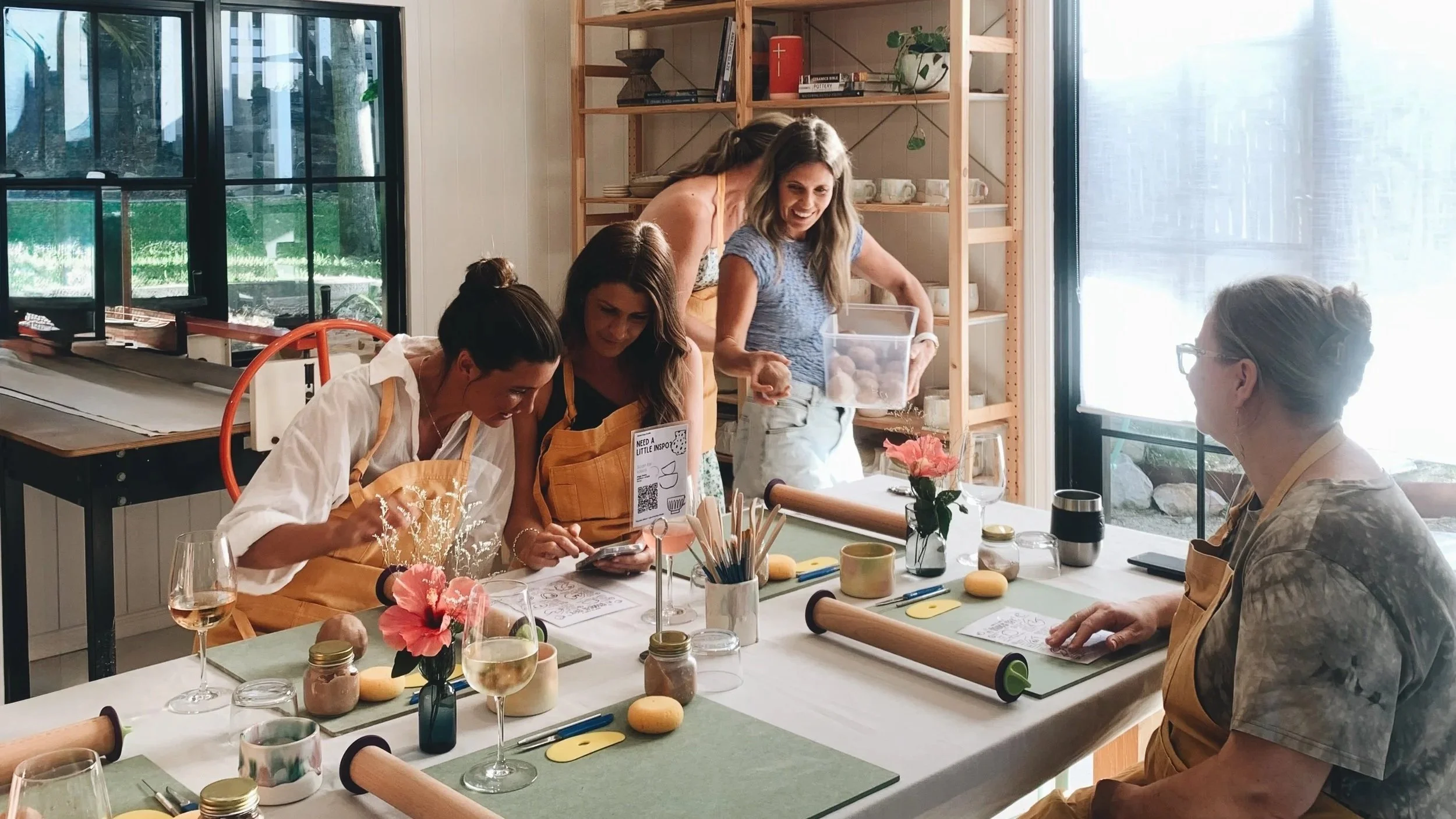 A group of women participating in a cooking or baking class around a table with rolling pins, plants, and pink flowers, in a well-lit room with large windows.