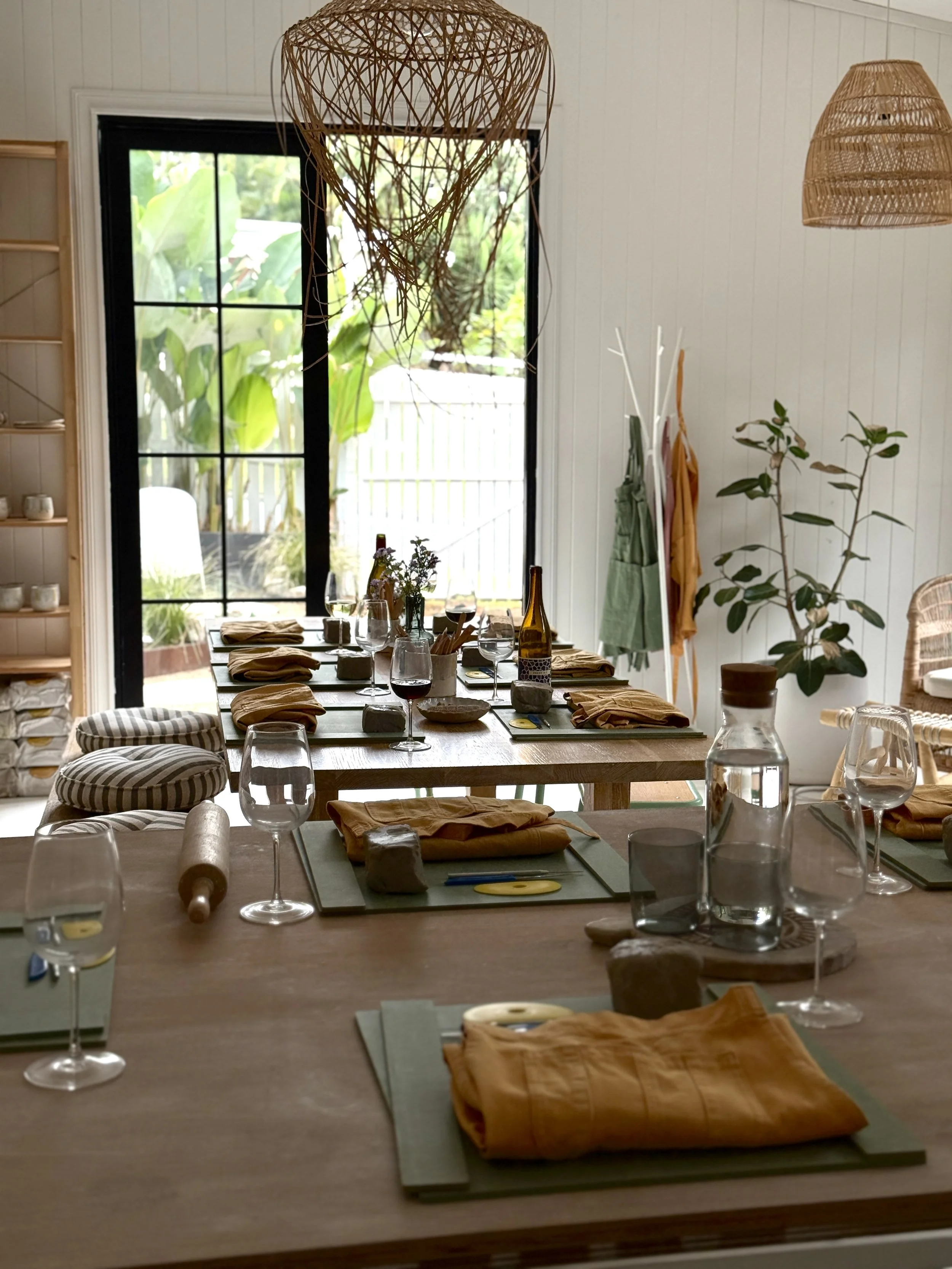 A dining table set for multiple people with rolled napkins, glasses, plates, and bottles, in a bright room with large windows, plants, and woven pendant lights.