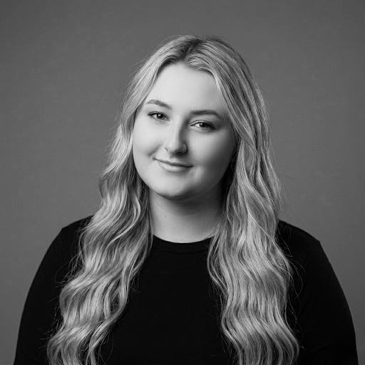 A young woman with blonde, wavy hair wearing a black shirt, smiling softly against a gray background.