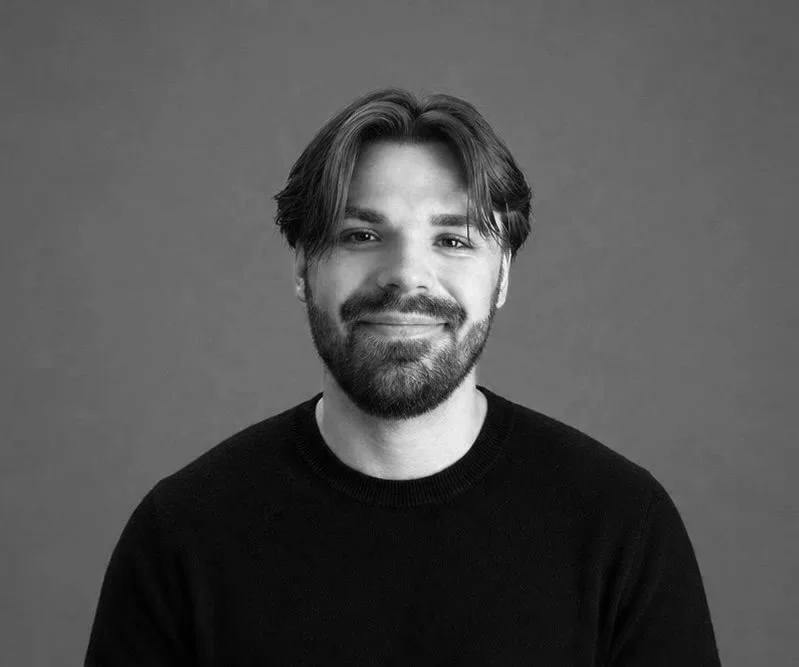 A smiling man with a beard and medium-length brown hair, wearing a black shirt, against a gray background.