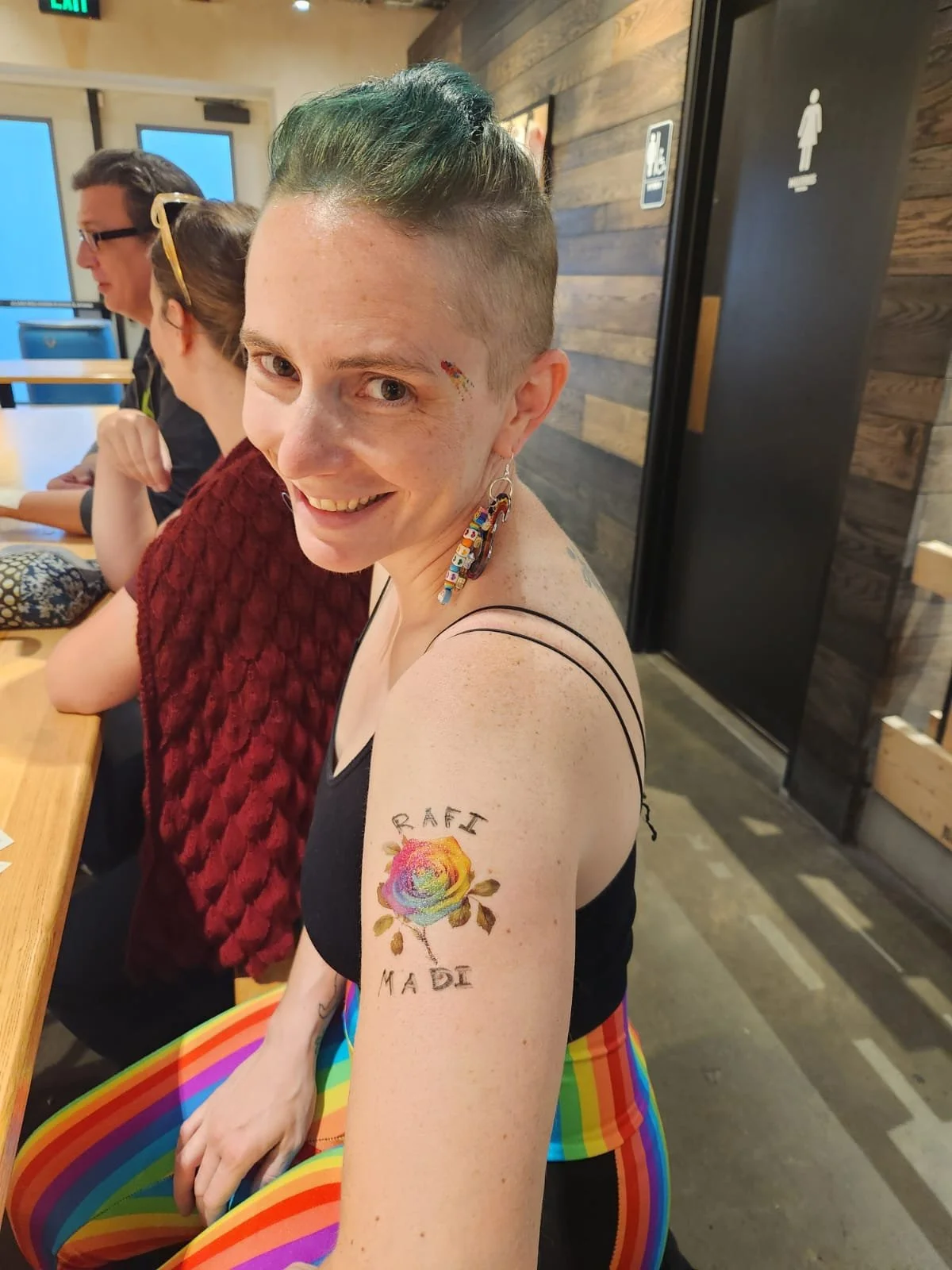 A smiling person with short blue hair, rainbow-colored earrings, and rainbow tattoo on her upper arm, sitting at a table with rainbow-striped pants in a casual indoor setting with wooden walls. She has a small rainbow sticker on her face.