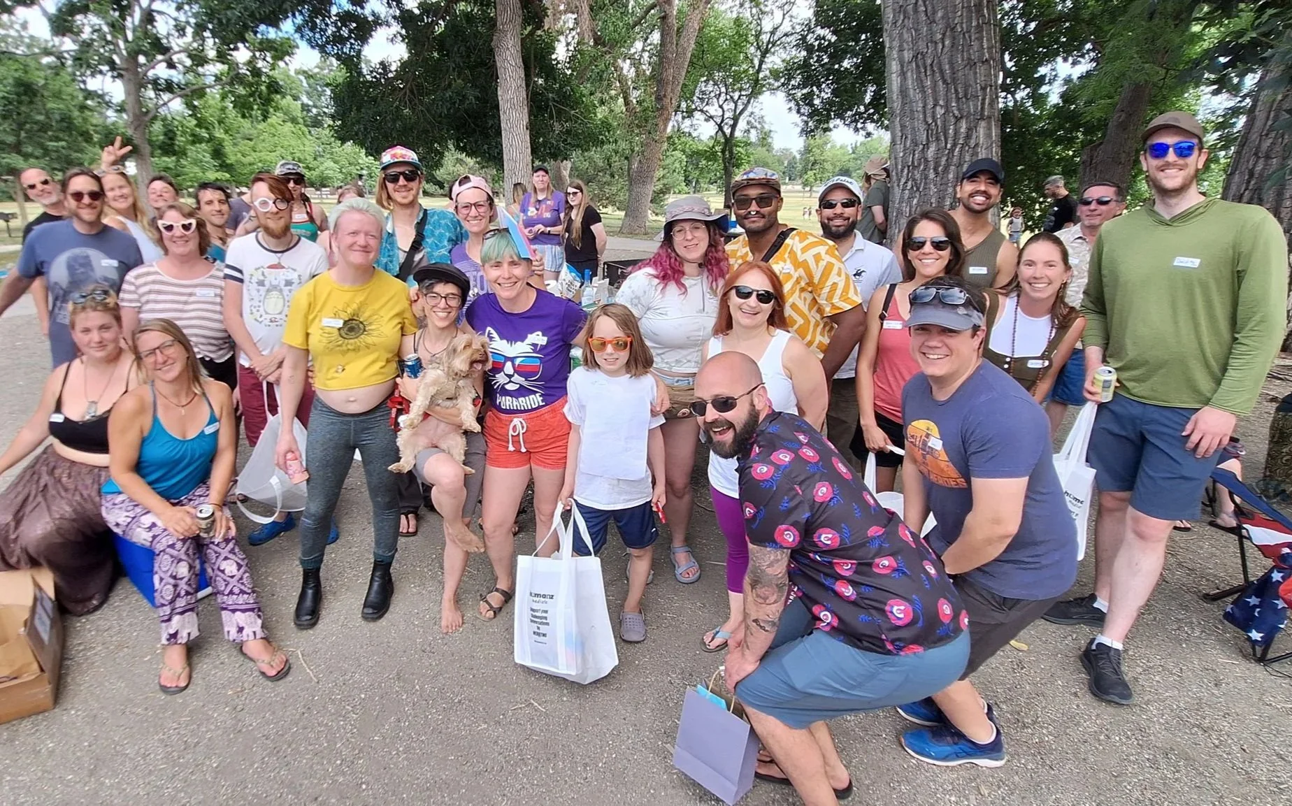 Group of diverse people gathered outdoors in a park, smiling at the camera, some holding bags and wearing casual summer clothes, with trees in the background.