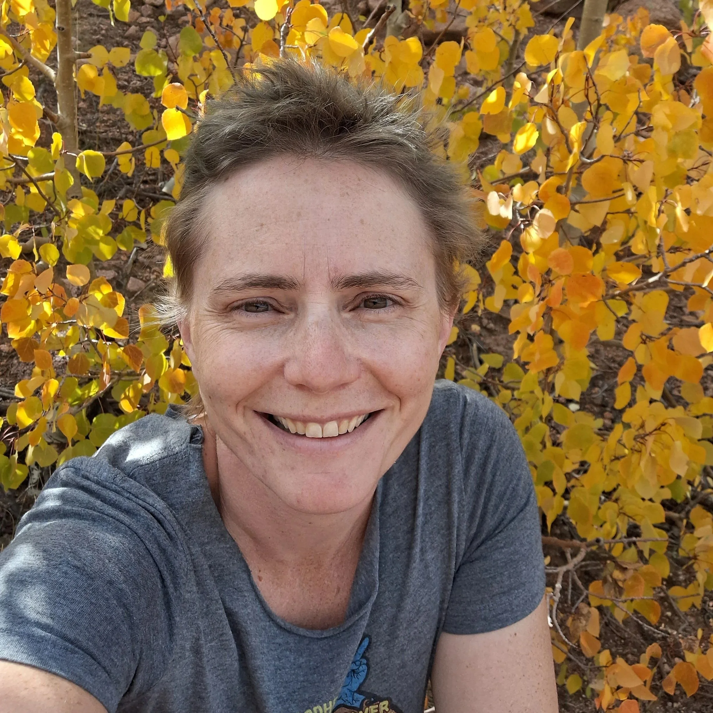 Close-up of a smiling person with light eyes and short hair, wearing a gray shirt with a graphic, outdoors in front of yellow-leaved shrub.