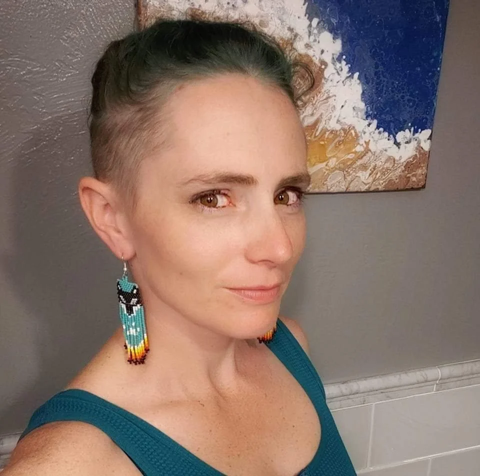 A person with short, blue hair styled up, wearing large beaded earrings with turquoise, black, and yellow colors, and a teal tank top, standing in front of a gray wall with a colorful abstract painting.