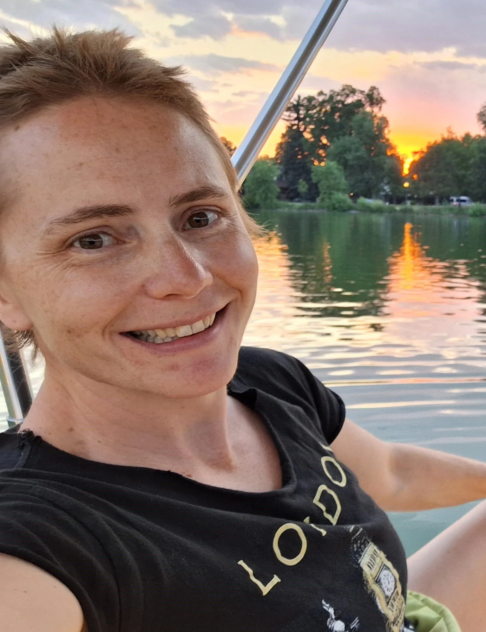 A person taking a selfie on a boat during sunset with trees and water in the background.