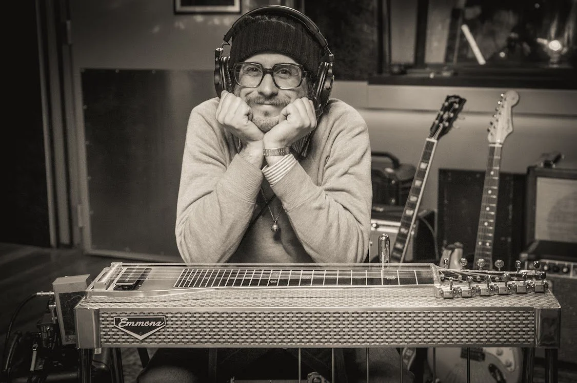 A man with glasses and a beanie sitting at a recording studio, resting his chin on his hands, with a lap steel guitar in front of him and other guitars in the background.