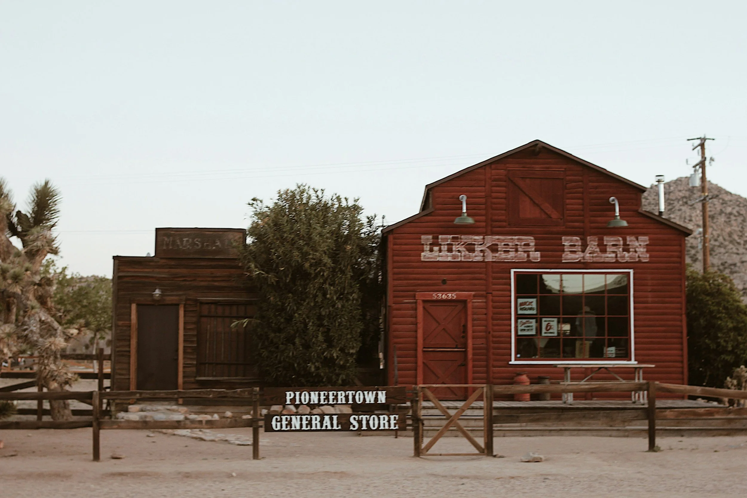 A red wooden building labeled 'Lucky Bar' with signs for a general store, located in Pioneer town, with a dirt road in front and desert plants around.