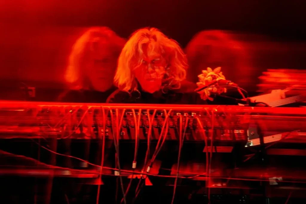 Steve Roach with blonde curly hair performing with electronic music equipment, illuminated by red lighting with a motion blur effect.