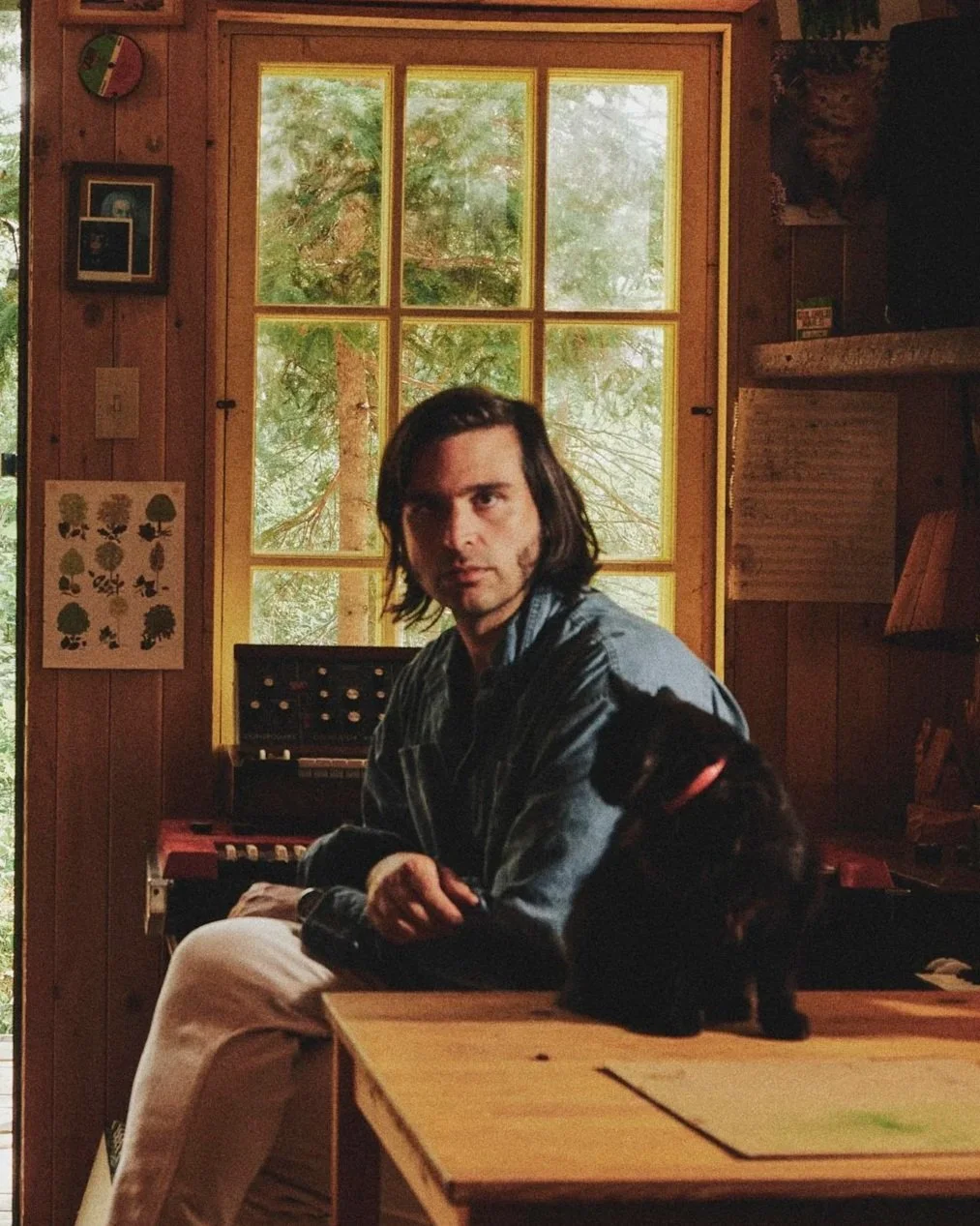 Flore Laurentienne with shoulder-length dark hair sitting at a wooden table inside a cozy room with wood-paneled walls and a large window showing green trees outside. A black dog with a red collar is sitting on the table beside him.