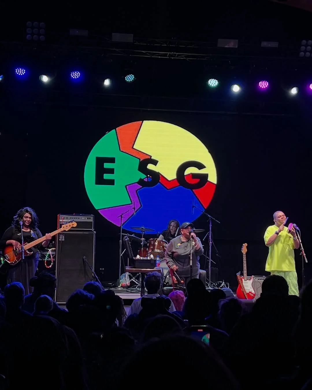 A live concert performance by  on stage. In the background, a colorful pie chart with the letters E, S, G, and a star is projected. The band members include a woman playing bass, a drummer, a woman singing with a microphone, and a man with a microphone in a bright yellow outfit. The audience is visible in the foreground.