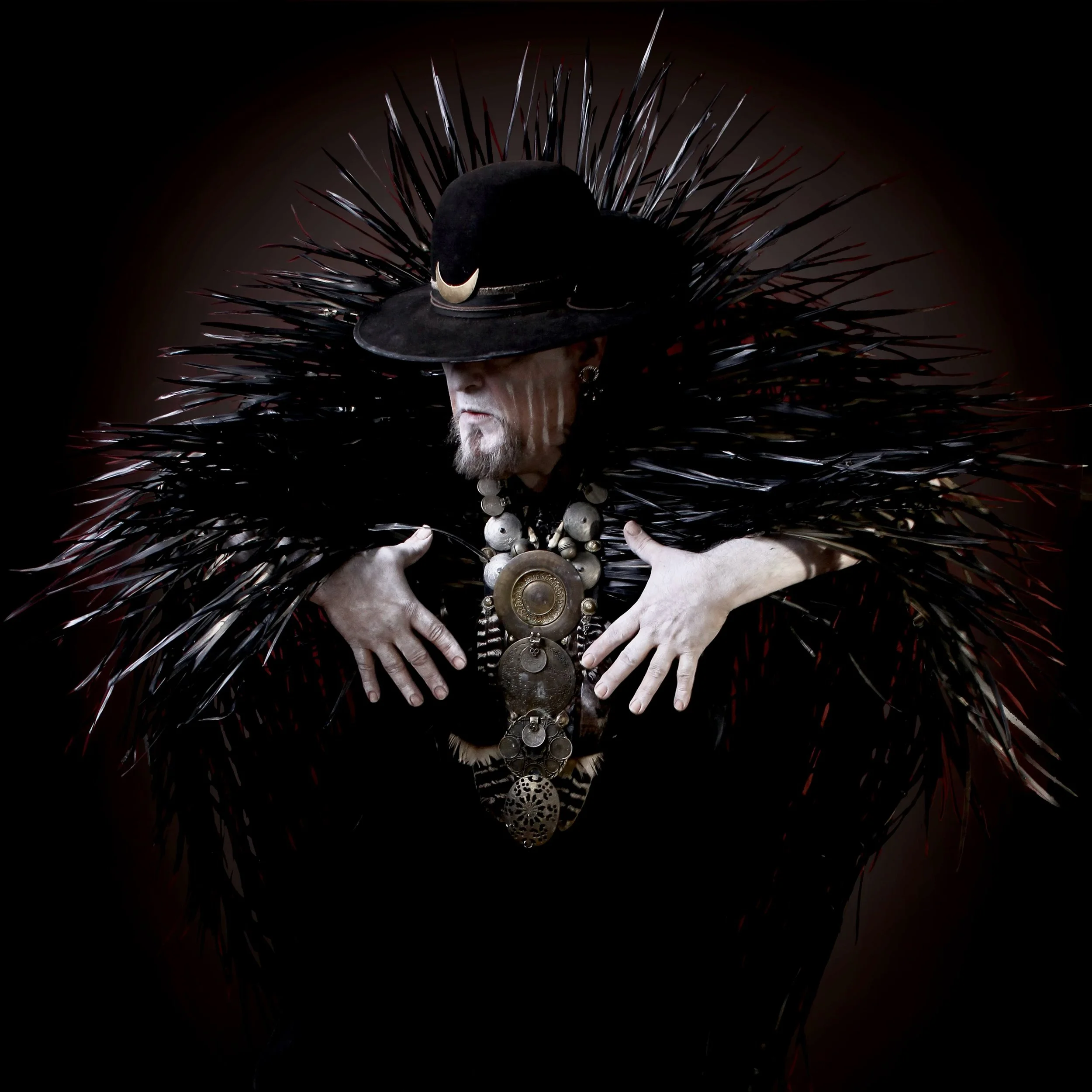 Person dressed in black with a large black hat adorned with metallic spikes and a large necklace, standing against a dark background.