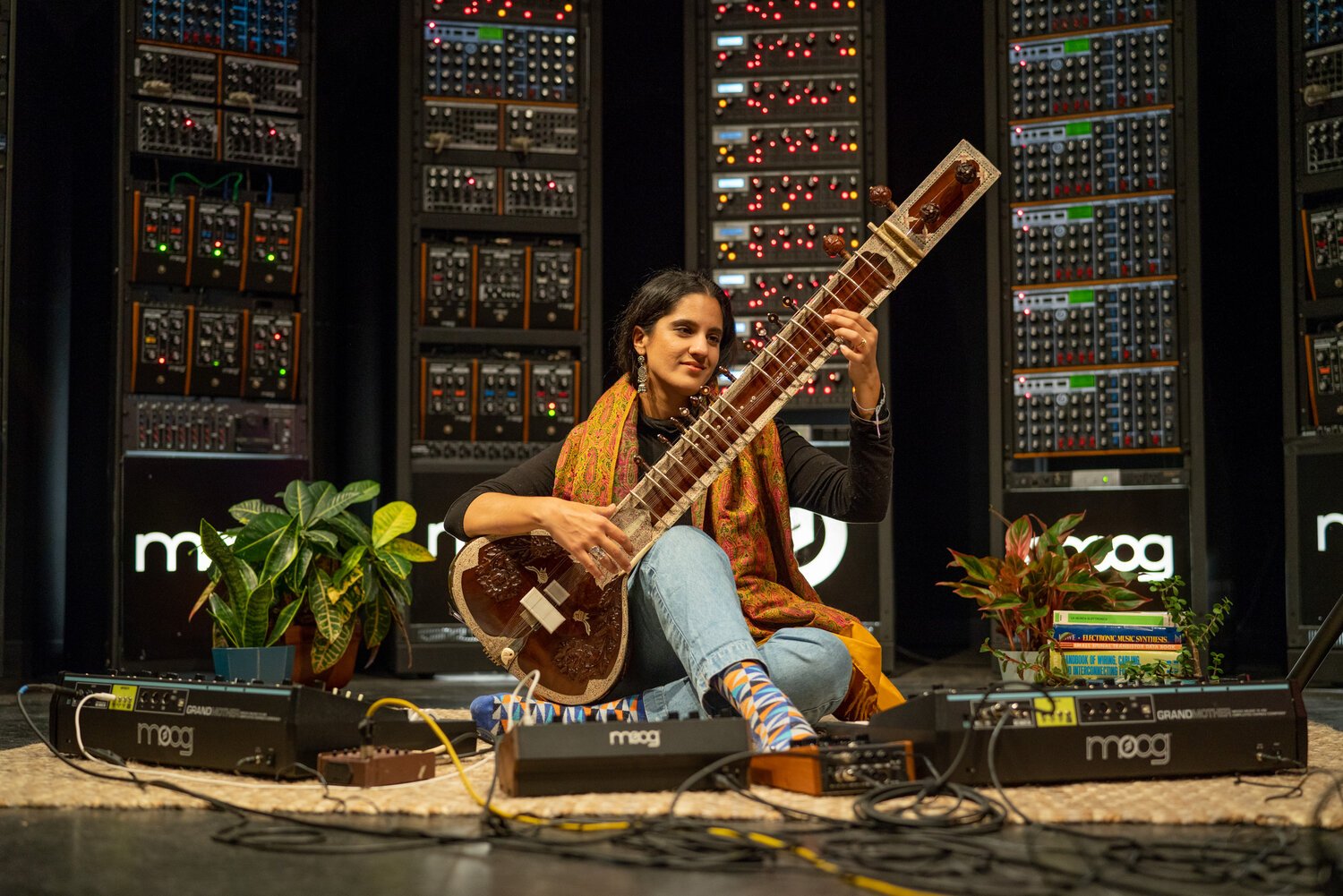Ami Dang, sitar musician on the floor in front of an array of electronic music equipment, playing a traditional instrument similar to a sitar, surrounded by potted plants and books.