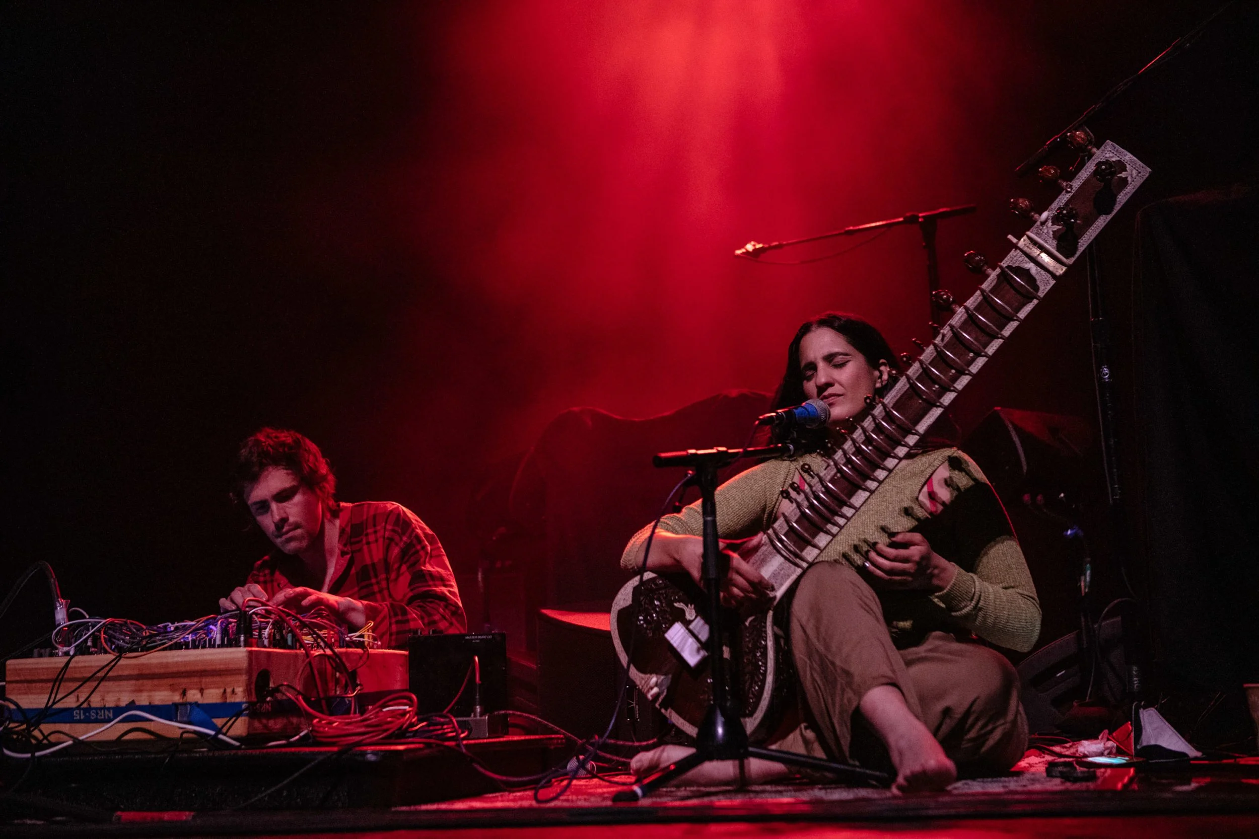Sitarist, Ami Dang performing on stage with red lighting, including a woman playing a sitar and a man working with electronic equipment.