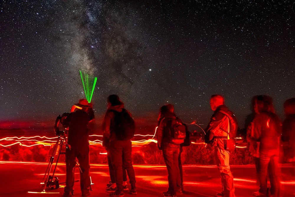 Group of scientists or astronomers observing the night sky with a telescope under a star-filled sky and the Milky Way galaxy visible.