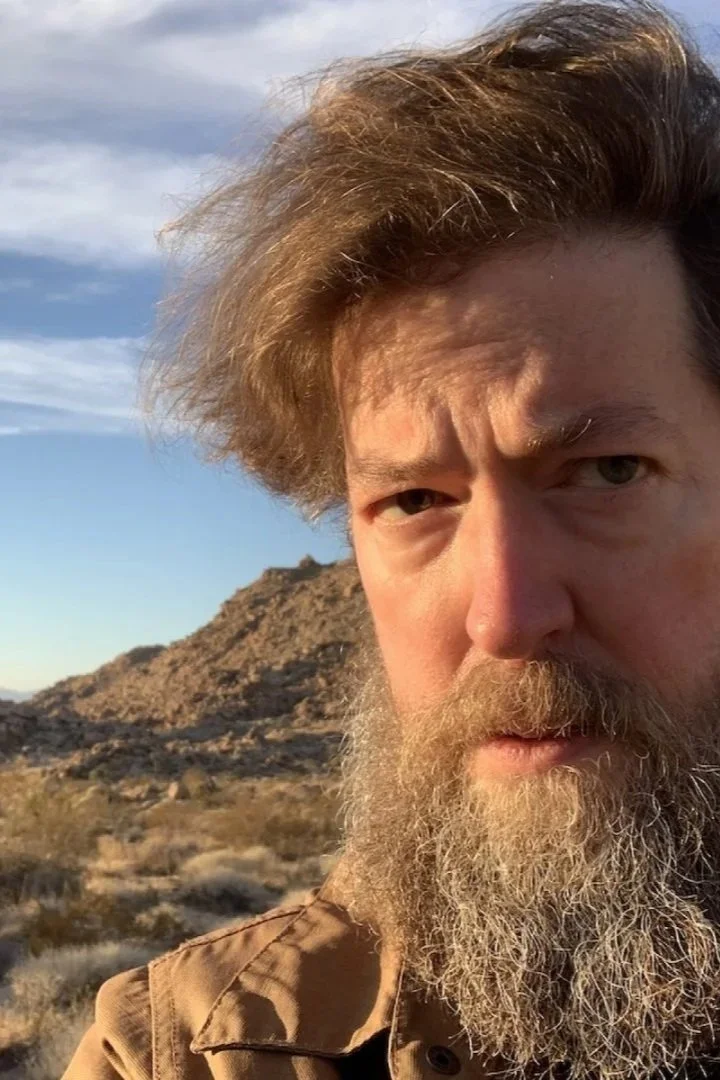 Ken Layne, host of Desert Oracle with a beard and tousled hair taking a selfie outdoors in a desert landscape with rocky hills and sparse desert vegetation, under a partly cloudy sky.