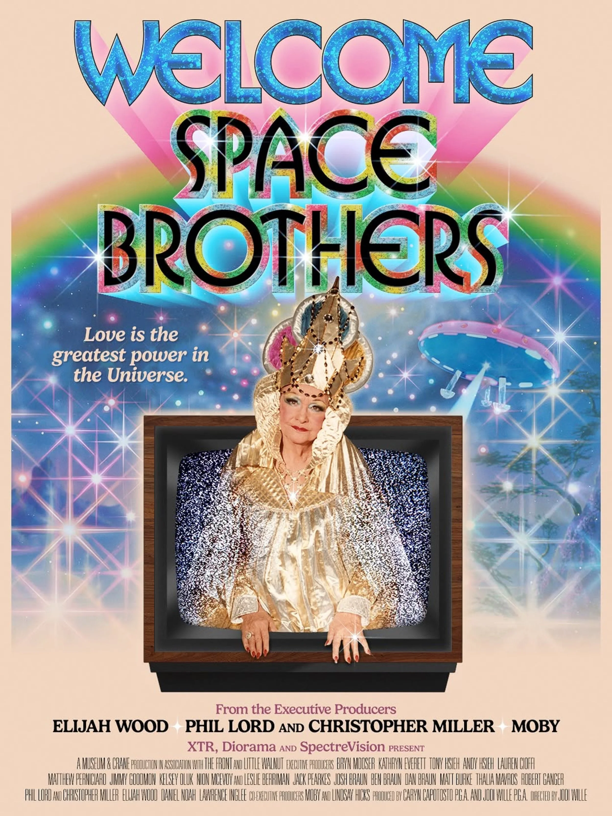 A colorful poster features the text 'Welcome Space Brothers' with a rainbow background, sparkling stars, and an image of a woman with elaborate gold and jeweled headwear emerging from a television screen.