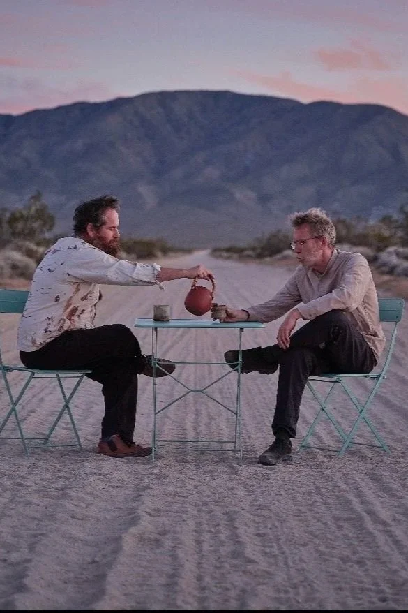 Two men sitting at a small table outdoors in a desert landscape with mountains in the background, sharing tea and cookies during sunset.
