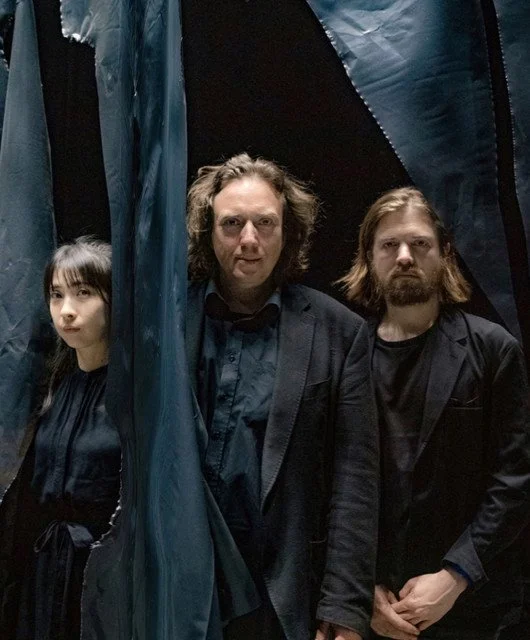 Tangerine Dream standing in a dark, tent-like environment, dressed in dark clothing, looking at the camera.