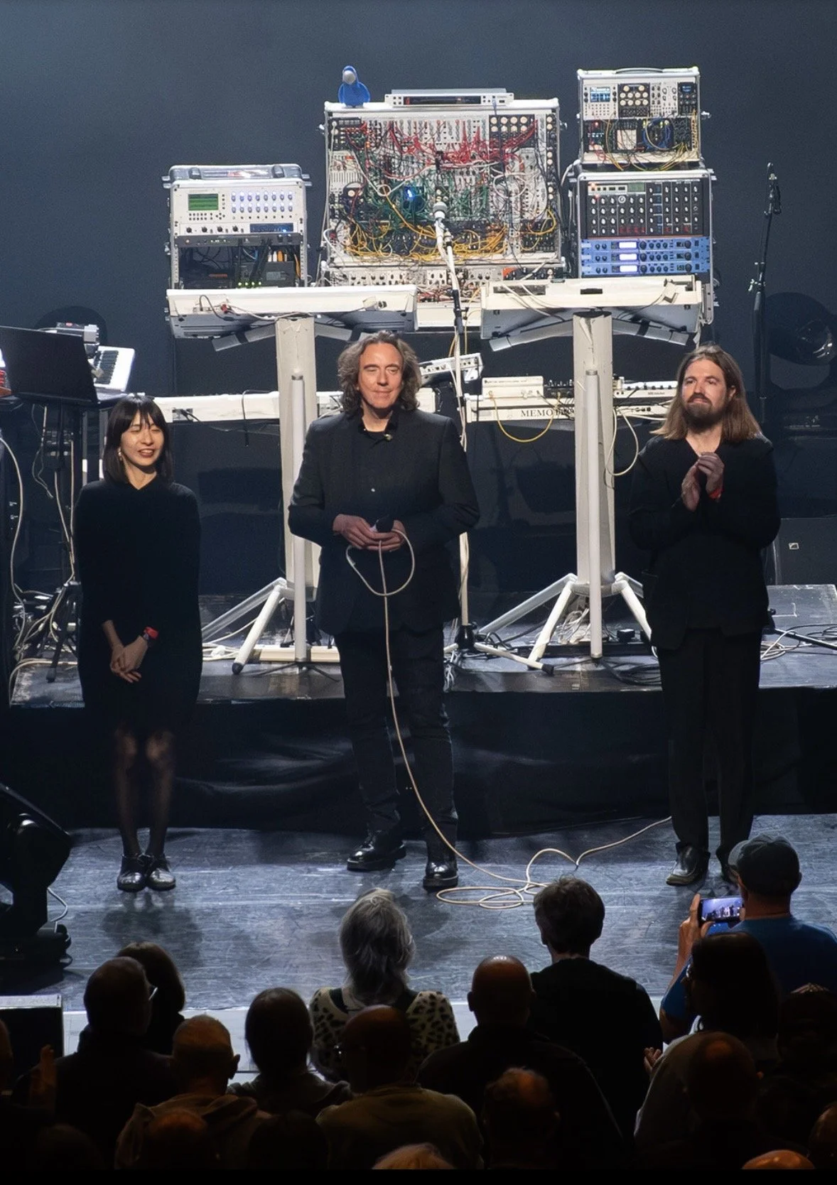 Tangerine Dream performers stand on a stage in front of an electronic music setup with modular synthesizers and equipment. The audience is watching, and one person is recording with a phone.