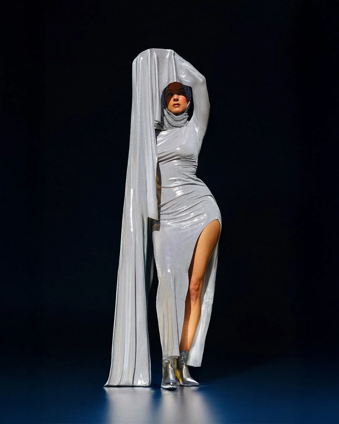 Alia theremin musician in a shiny silver dress with a high slit, wearing a matching hooded cape and silver high-heeled boots, standing against a dark background.