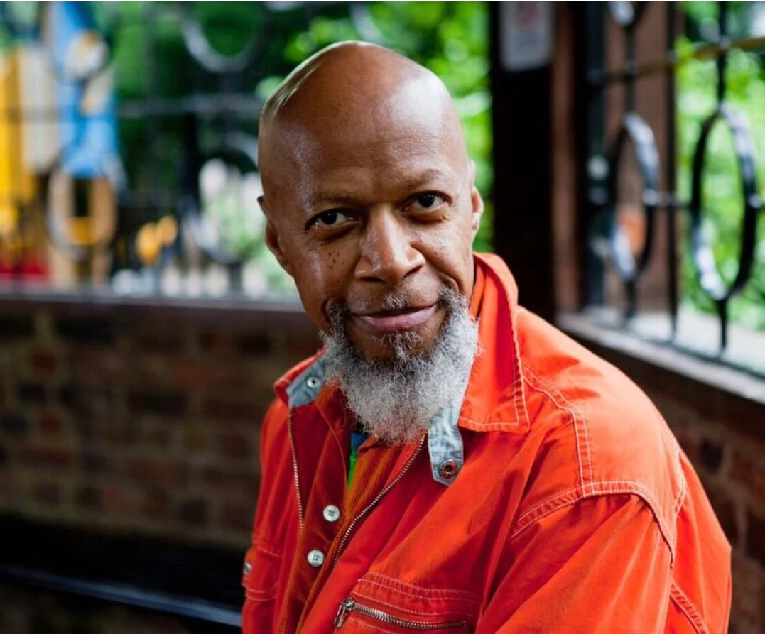 Laraahi with a bald head and a gray beard wearing an orange jacket, sitting outdoors with trees and a brick wall in the background.