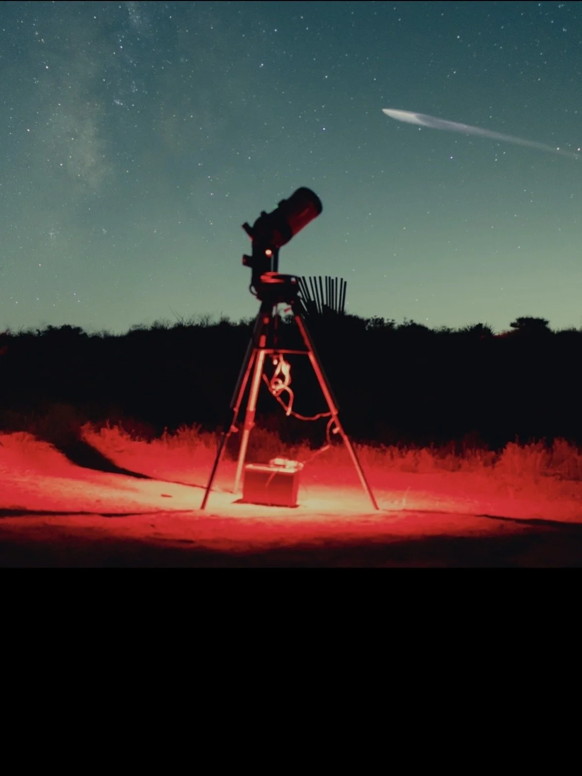 Night sky with stars and Milky Way galaxy, with shooting star visible. In foreground, a telescope on a tripod and a small red light source illuminating the ground near it.