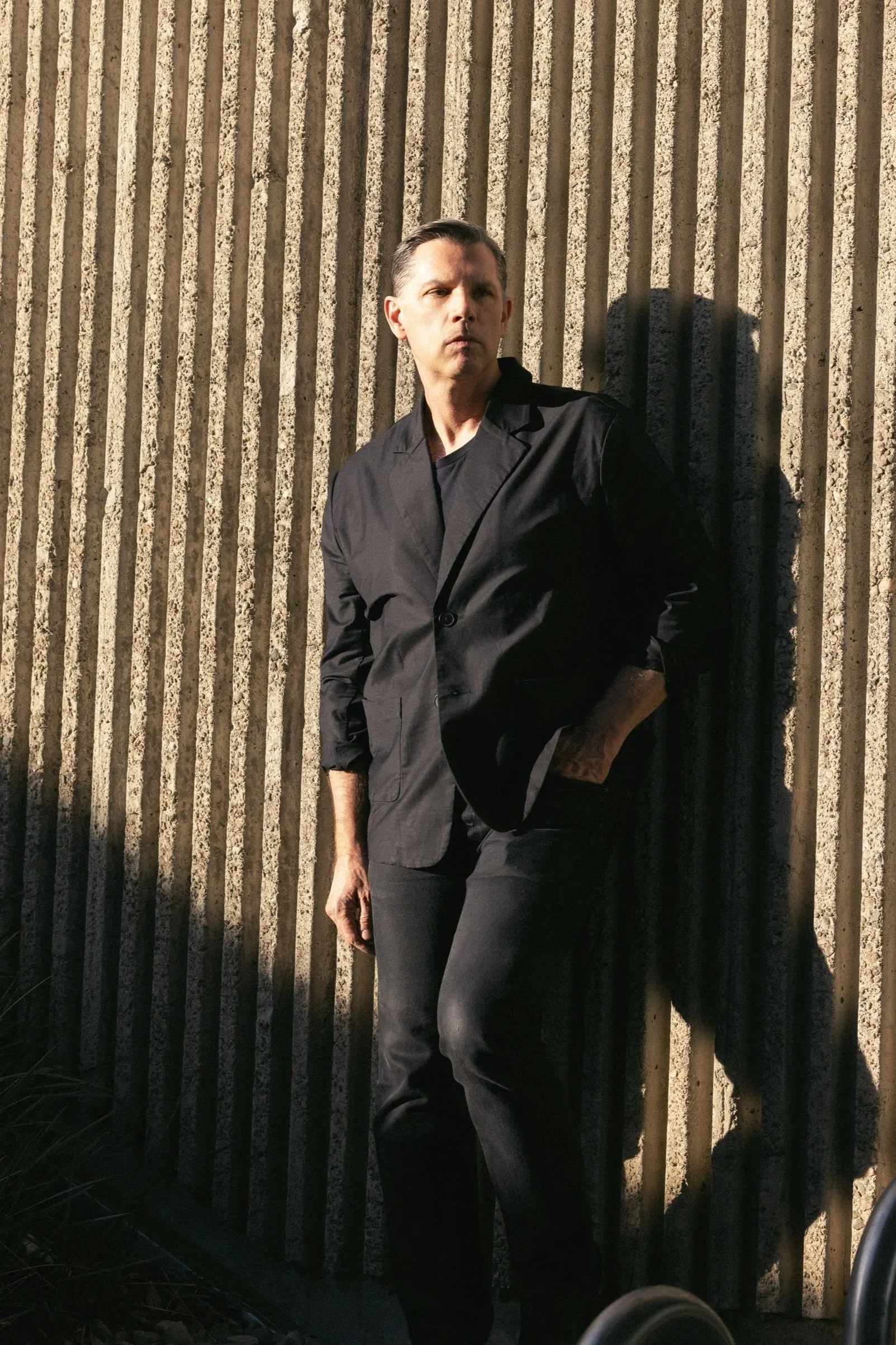 Daniel Paul  standing against a textured concrete wall in sunlight, with one hand in his pocket and looking to the side.