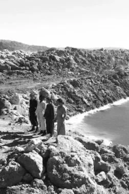 Four children standing on large rocks near a body of water, with a rugged landscape in the background.