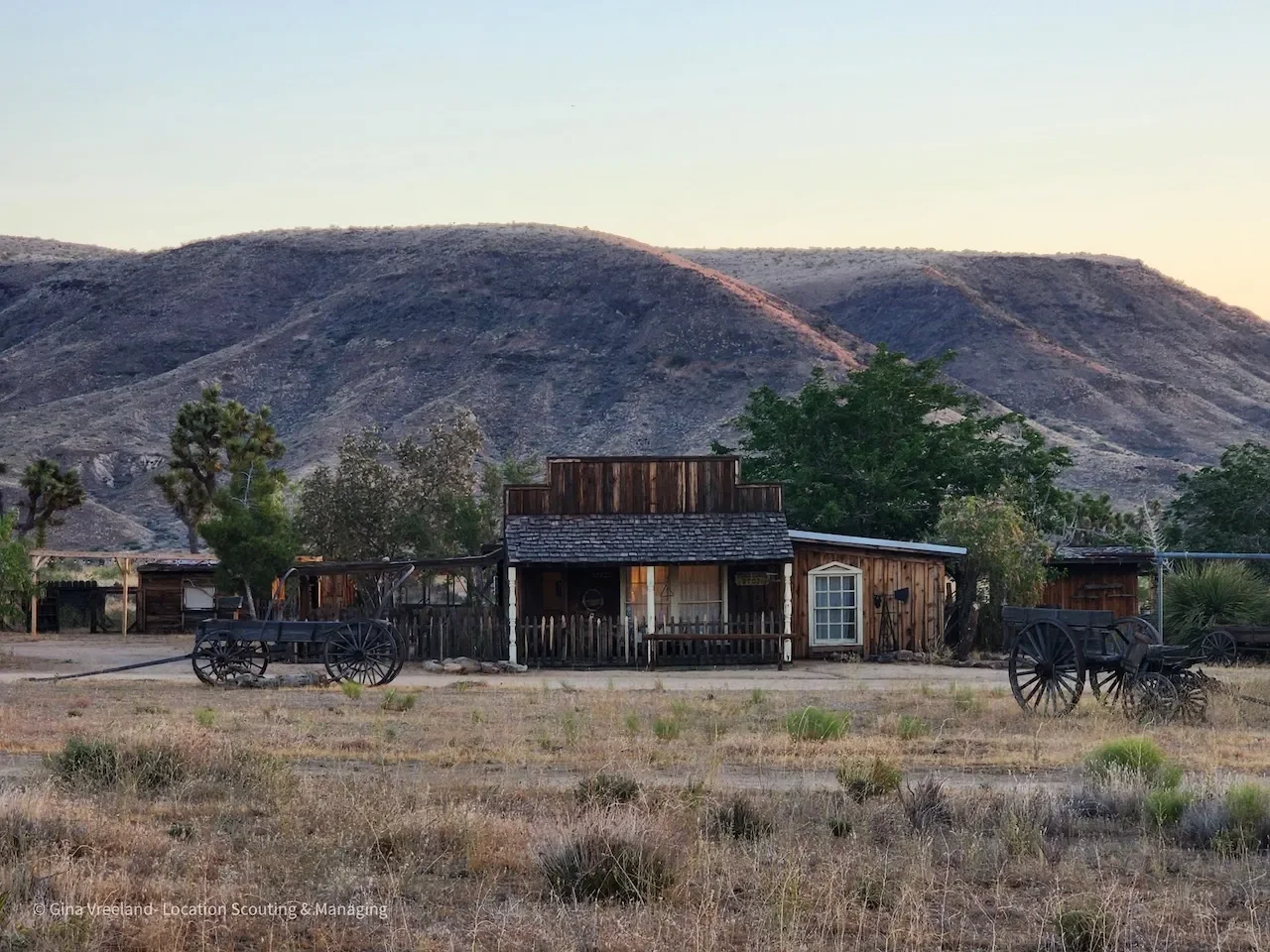 An old wooden house situated in a dry, grassy field with wagon wheels nearby and mountains in the background.